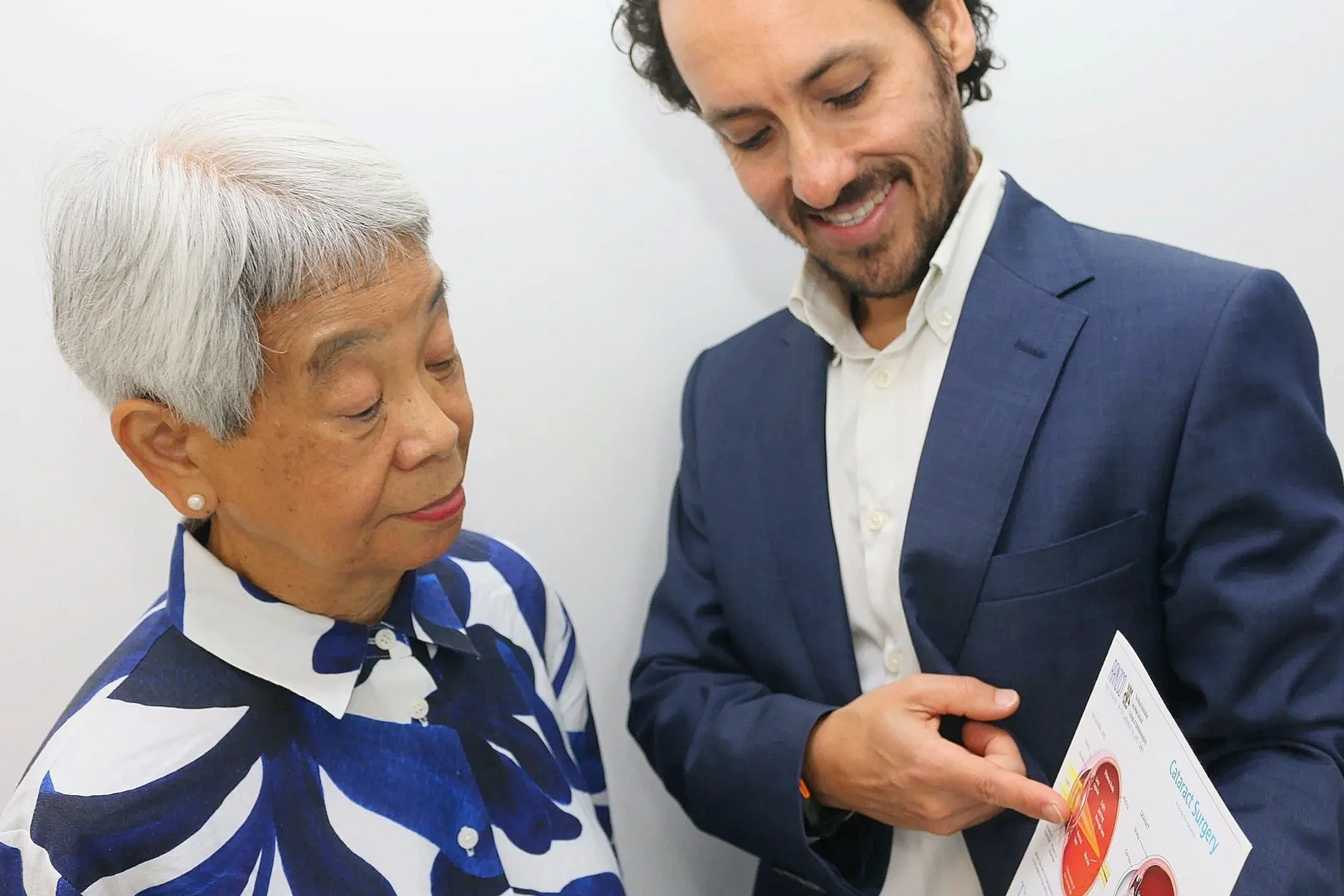 An elderly Asian woman with short white hair and pearl earrings wearing a blue and white patterned shirt looks at a pamphlet with a young man in a blue suit and white shirt, holding the pamphlet and smiling.