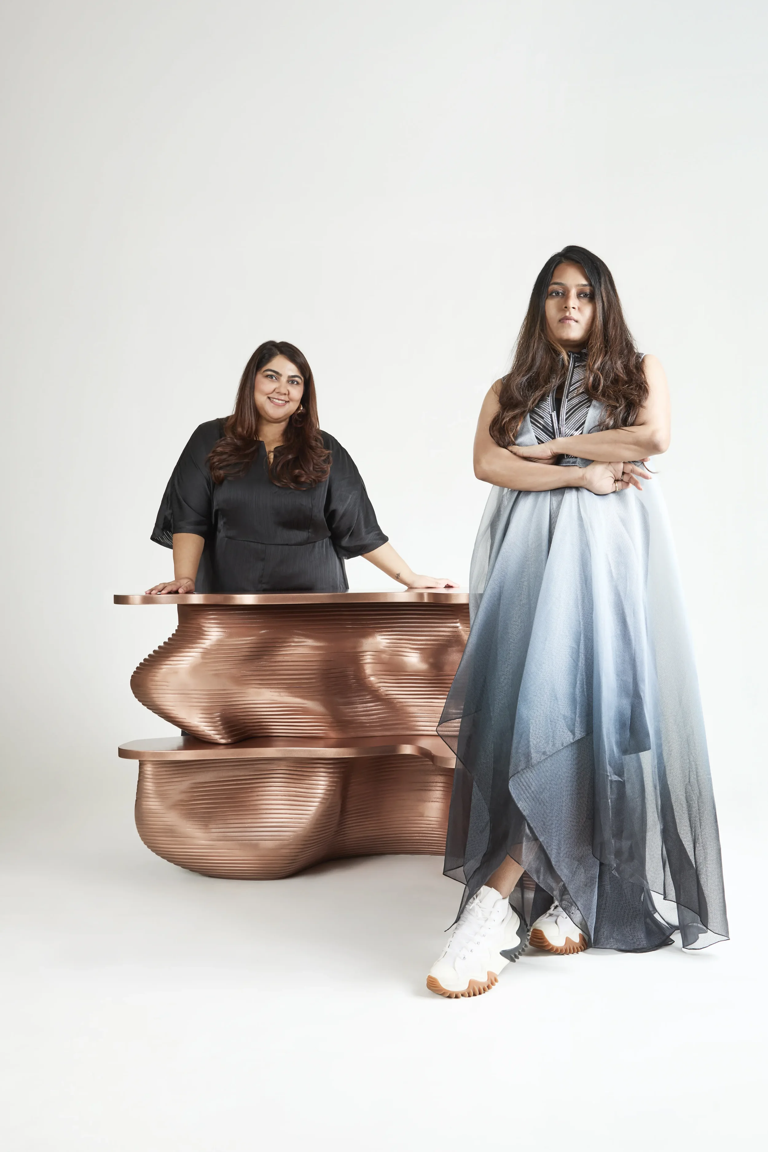 Two women in fashionable outfits pose against a plain white background, with one standing in front and the other behind a unique copper-colored sculptural table.