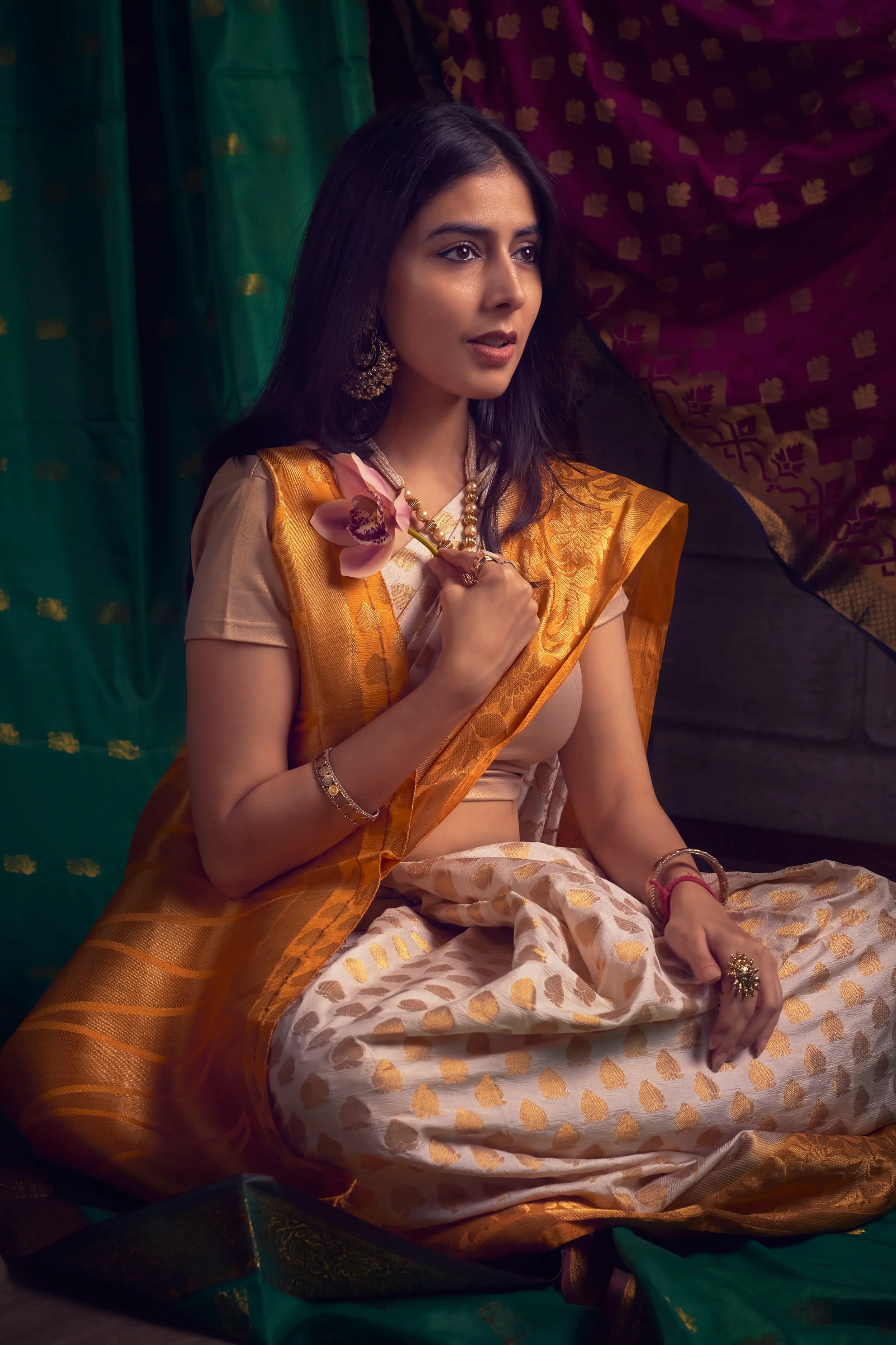 Woman dressed in traditional Indian saree, sitting cross-legged with a flower and jewelry, surrounded by colorful fabrics.