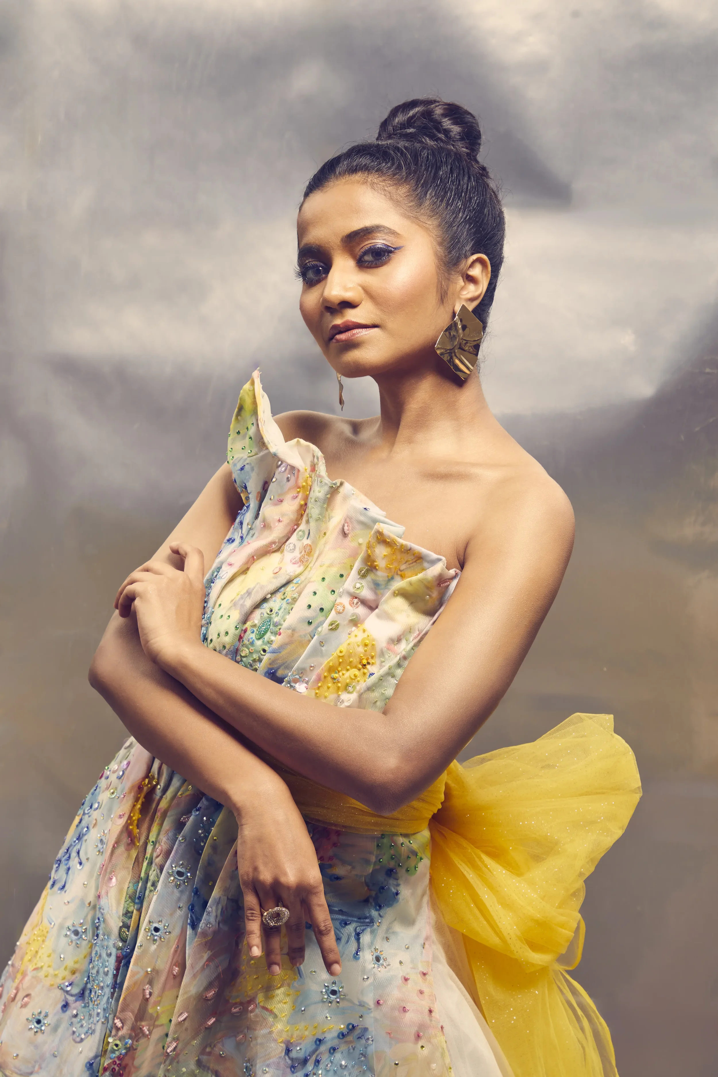 A woman dressed in a colorful, embroidered gown with a large yellow bow on the back, posing with folded arms against a neutral background.