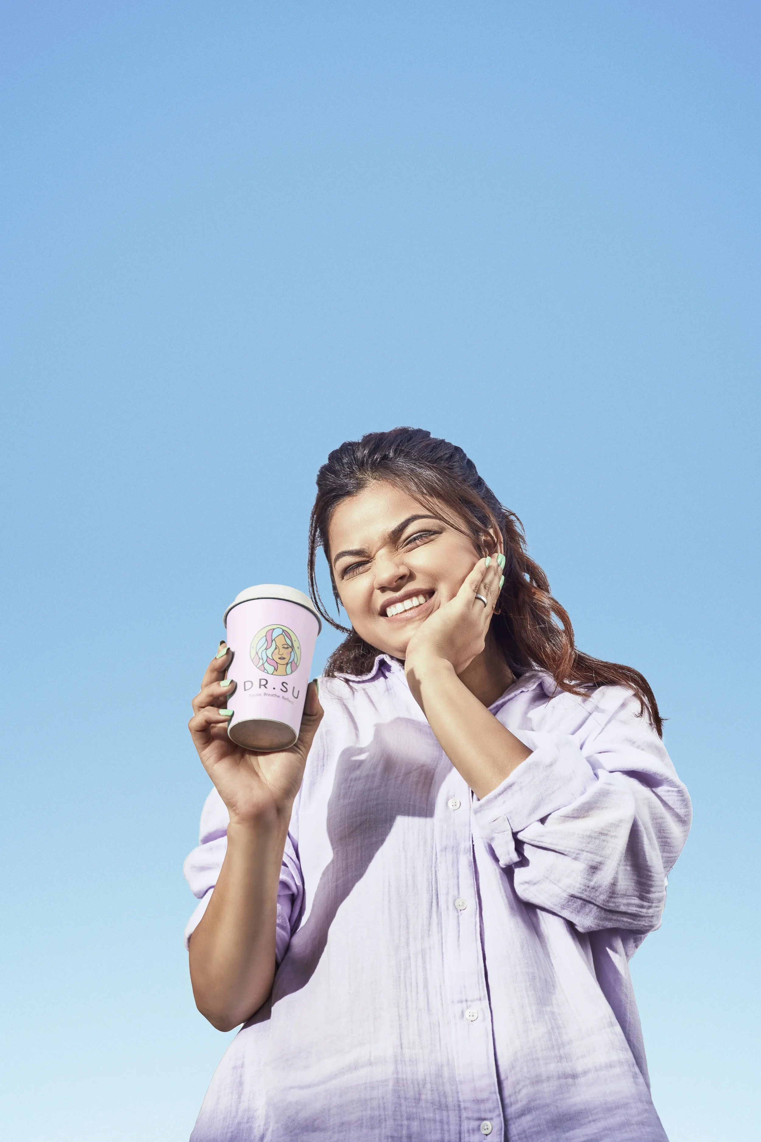 A woman with long brown hair, wearing a light purple shirt, smiling and holding a pink coffee cup with a logo, standing against a bright blue sky.