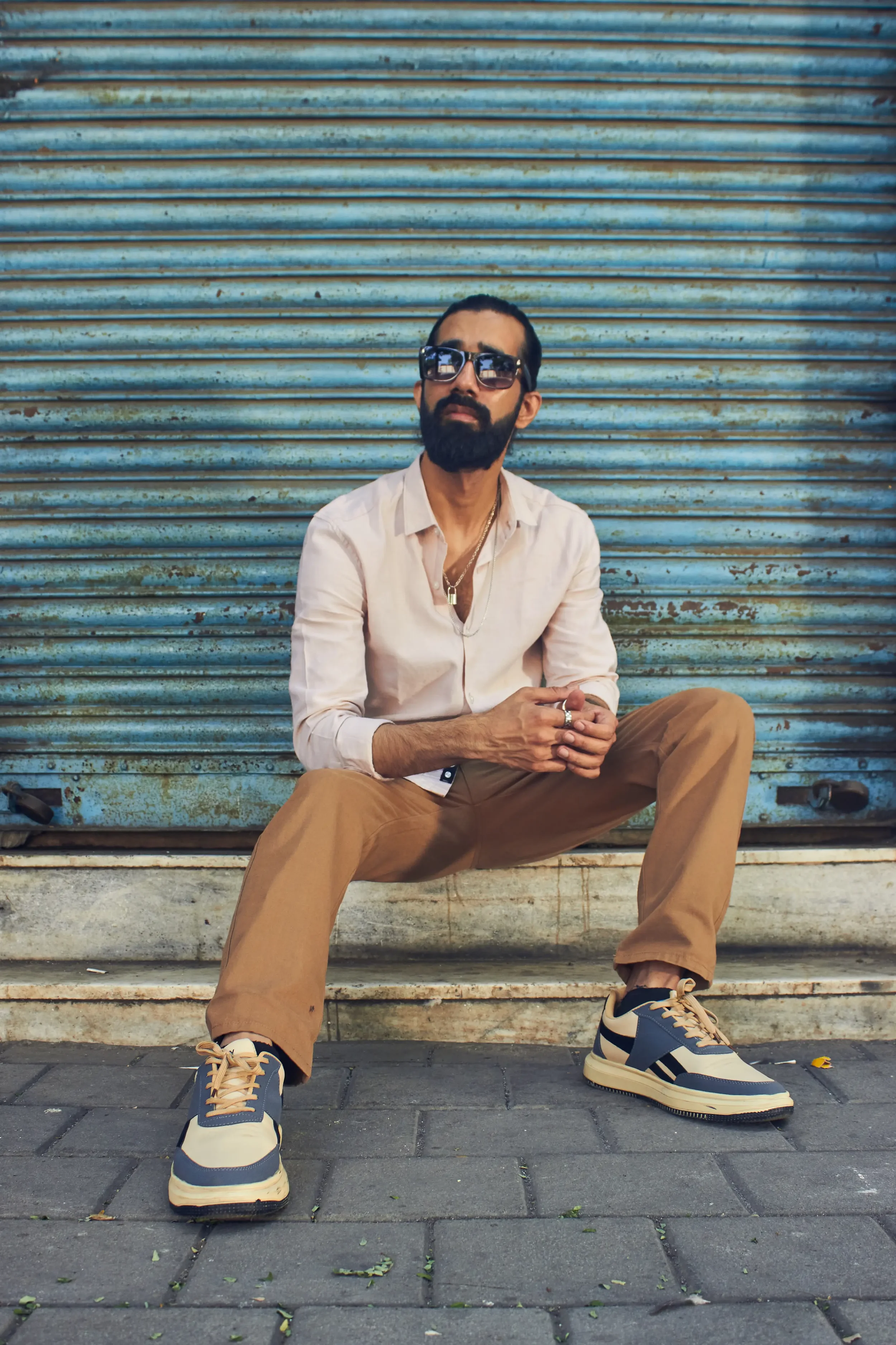 A man with a beard and sunglasses sitting on steps in front of a blue, rusty metal shutter.