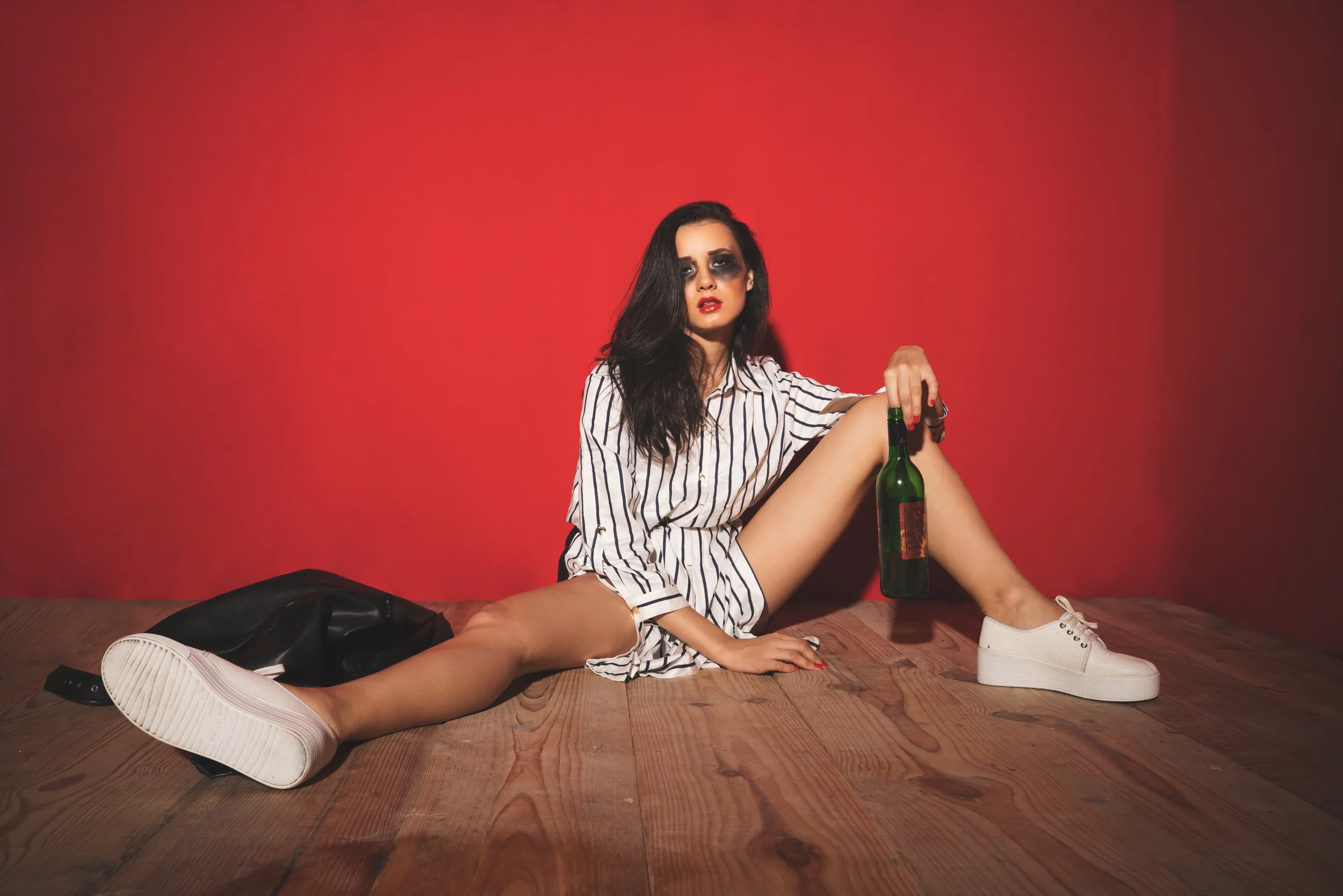A woman with dark hair and makeup sitting on a wooden floor, wearing a striped shirt and white sneakers, holding a green alcohol bottle, against a red wall.