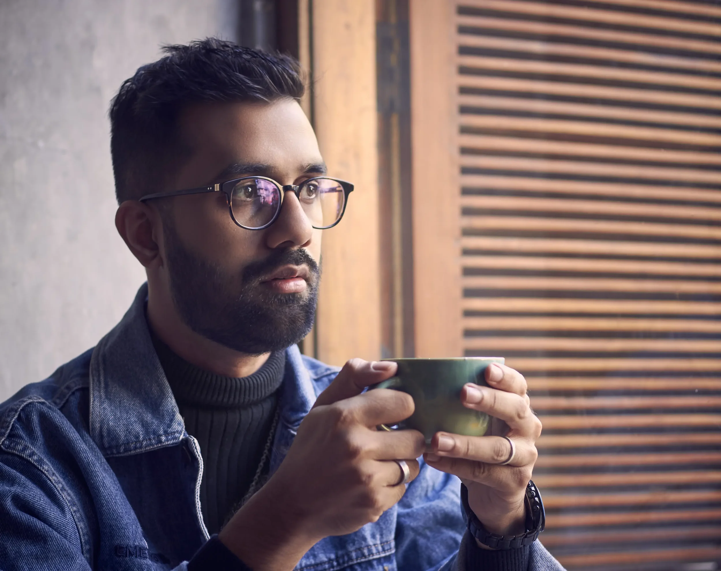 A man with dark hair, glasses, and a beard holding a greenish ceramic mug, sitting near a window with wooden blinds.