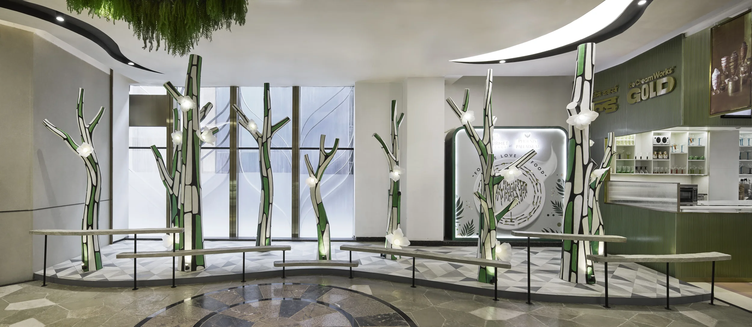 Interior of a modern cafe or food court with decorative illuminated tree-like structures in green, black, and white. There are benches in front of the trees, and a counter with shelves and a microwave on the right side.