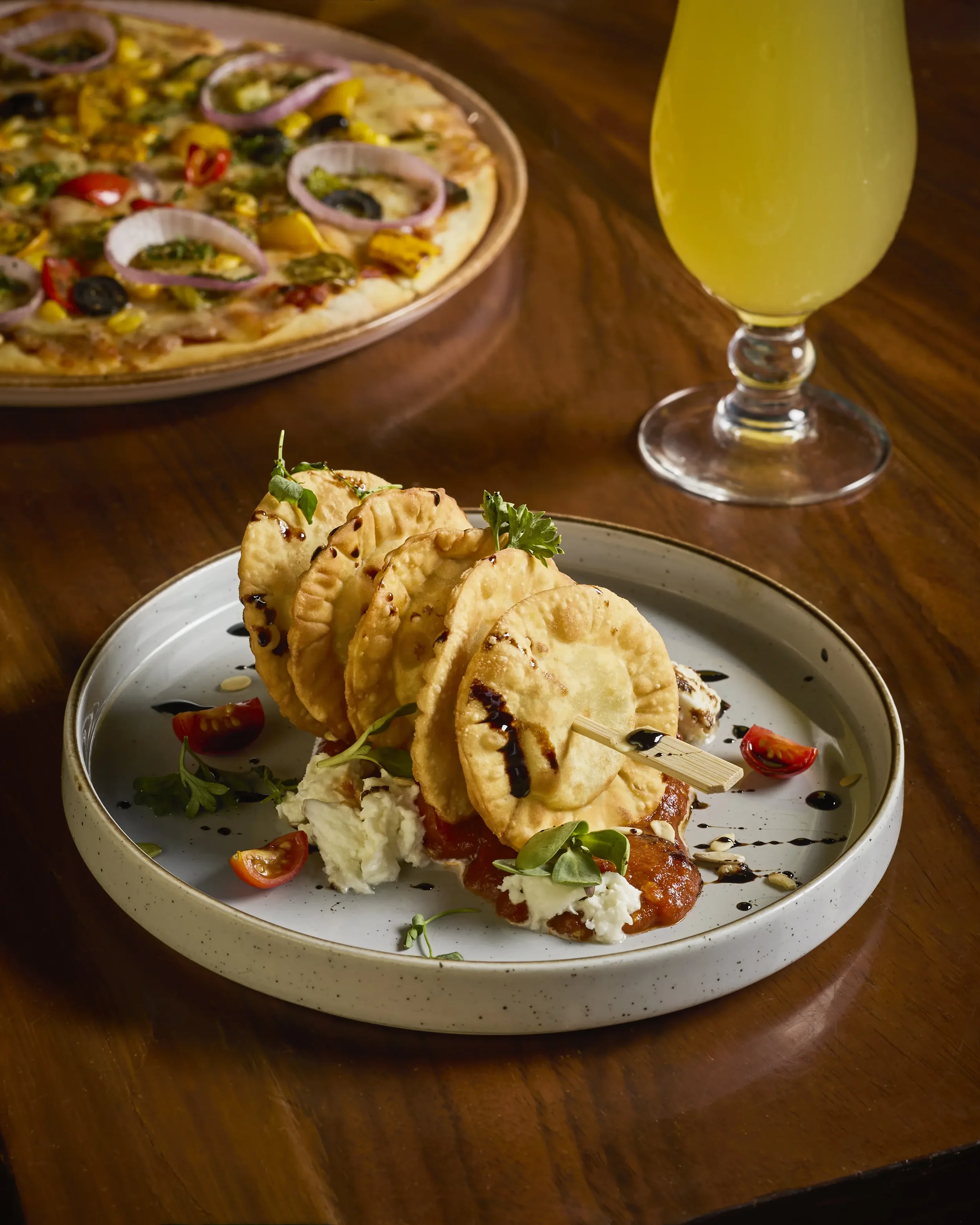 A plate of fried calamari garnished with herbs and cherry tomatoes, accompanied by mashed potatoes and drizzled with balsamic glaze, with a pizza and a yellow beverage in the background on a wooden table.