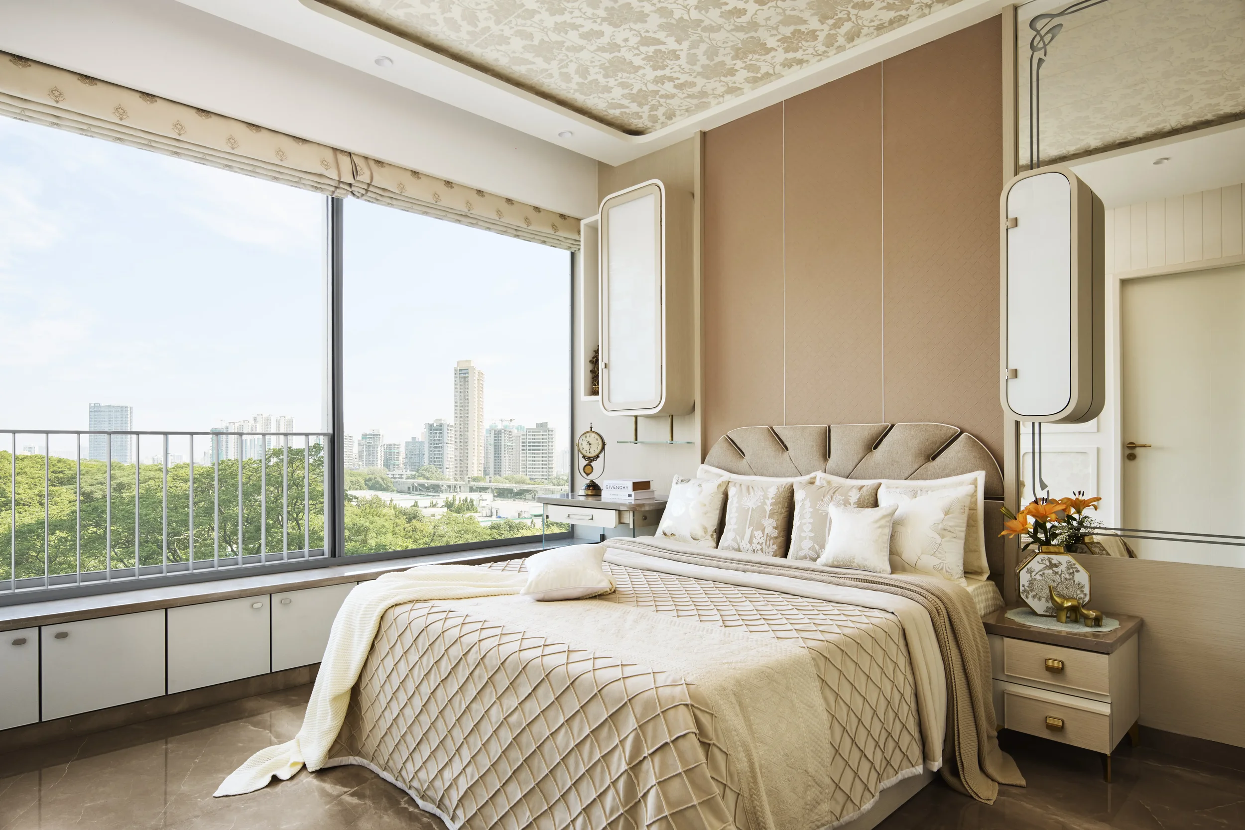 A bedroom with a large window showing city buildings and greenery outside. The bed has a beige upholstered headboard, multiple white pillows, and a cream-colored quilt. There are side tables with decor and a view of the city skyline.