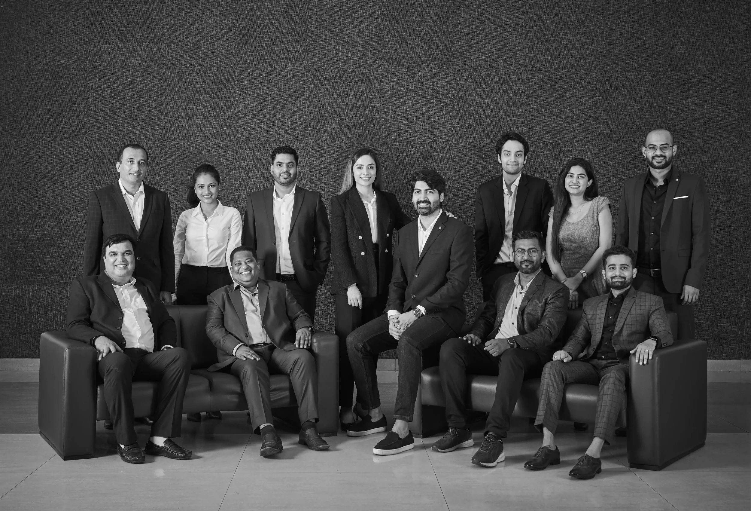 Group of fifteen people, dressed in business attire, posing in an office or conference room setting, some seated on sofa chairs and others standing behind, smiling at the camera.