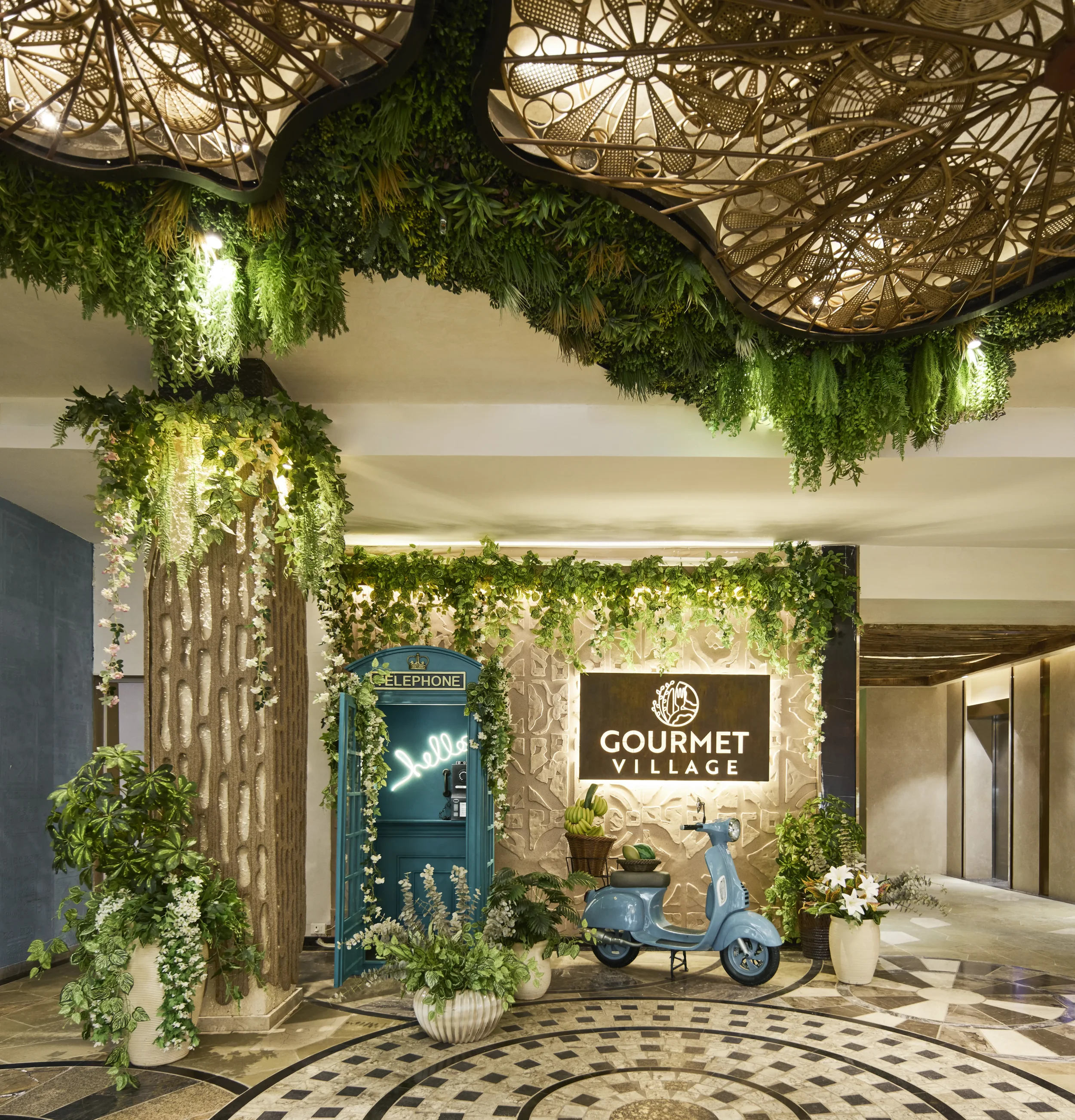 Decorative indoor space with lush green plants, a turquoise children's ride-on vehicle, and signs for 'Gourmet Village' and 'Telephone,' featuring natural and artistic design elements.