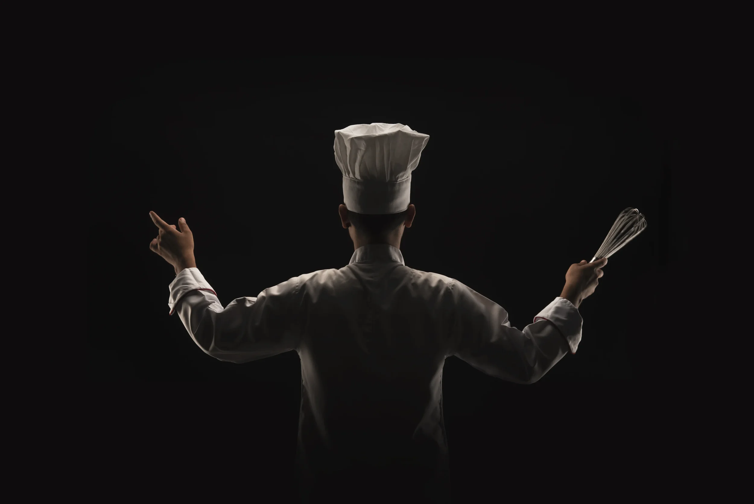 Silhouette of a chef wearing a tall white chef hat and coat, holding a whisk in one hand, against a dark background.