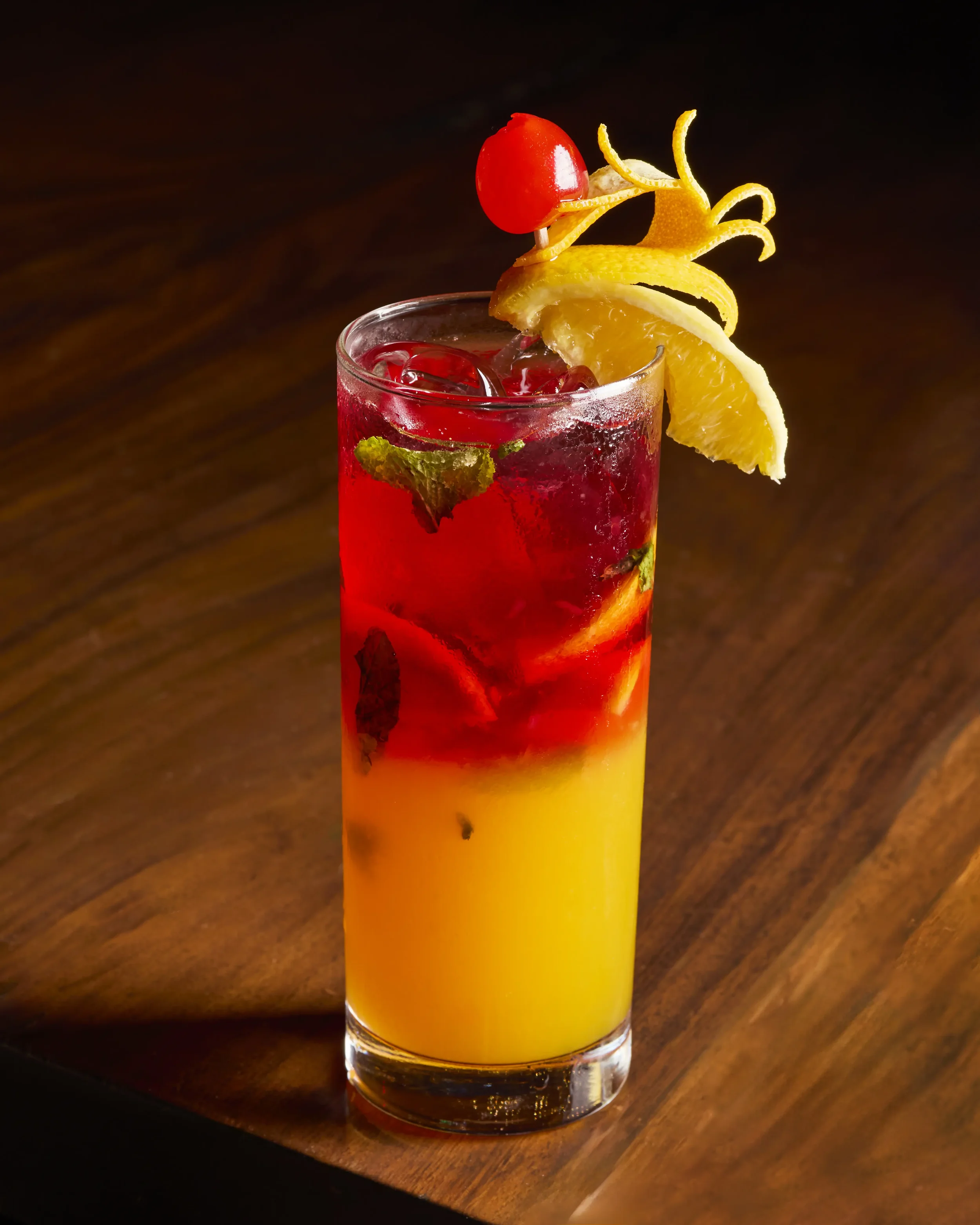 A colorful cocktail garnished with a cherry, lemon, and orange peel on a dark background.