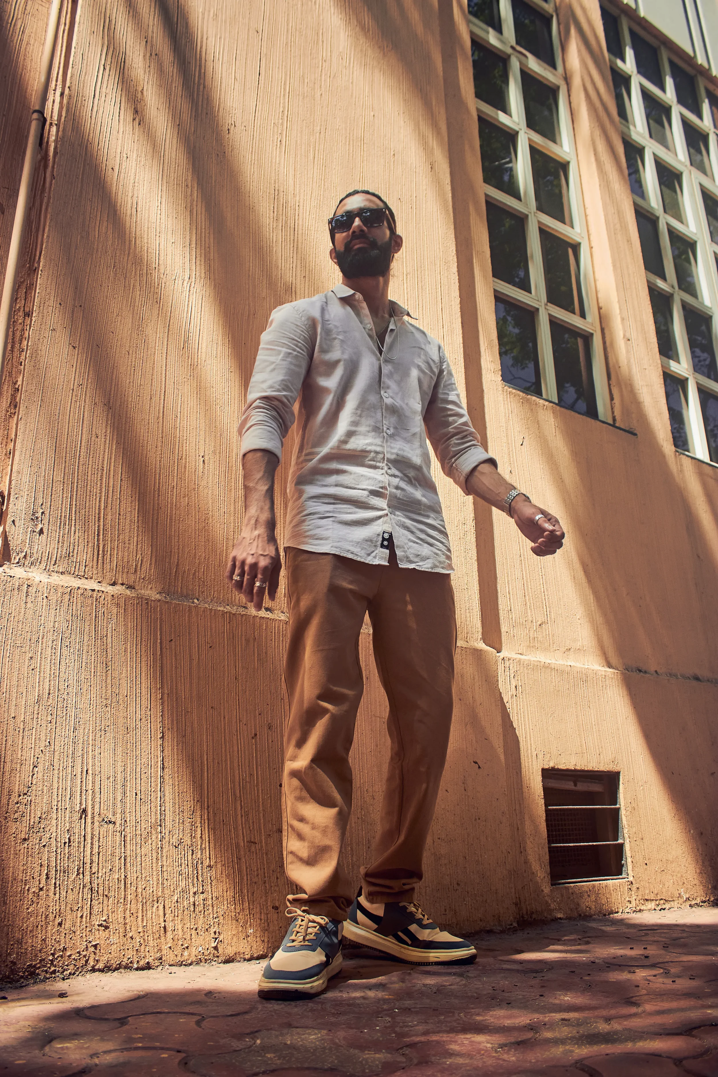A man wearing sunglasses, a light-colored shirt, beige pants, and sneakers standing outdoors against a textured pink wall with large windows.