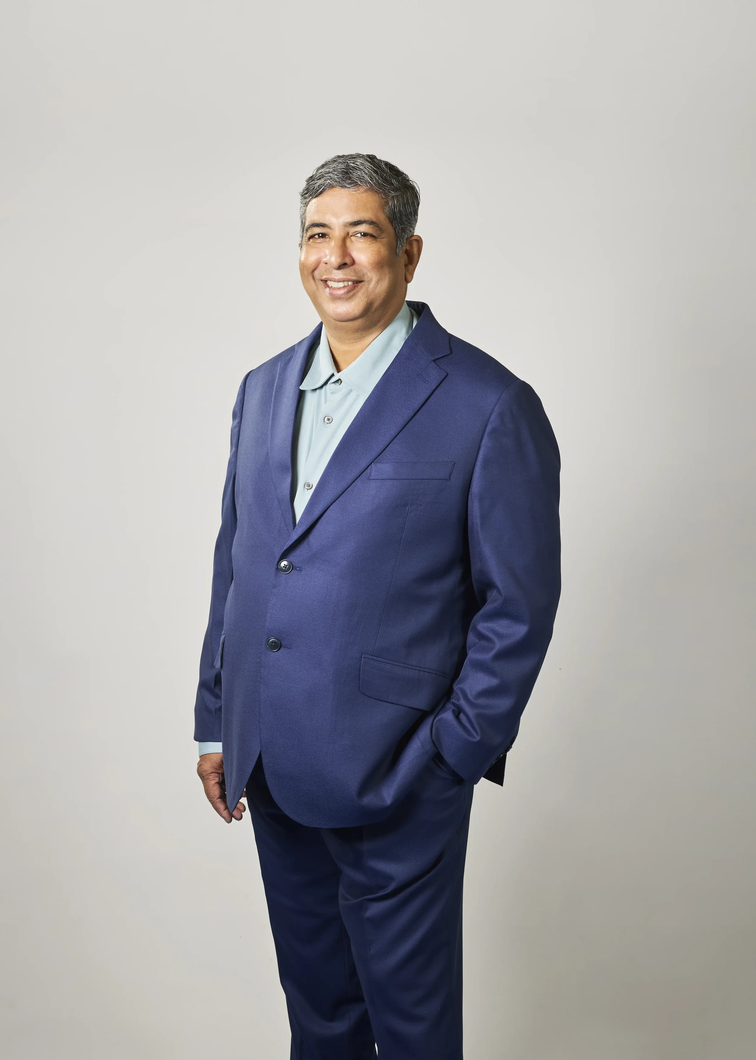 A middle-aged man with short gray hair wearing a blue suit and light blue shirt, smiling and standing in front of a plain light gray background.