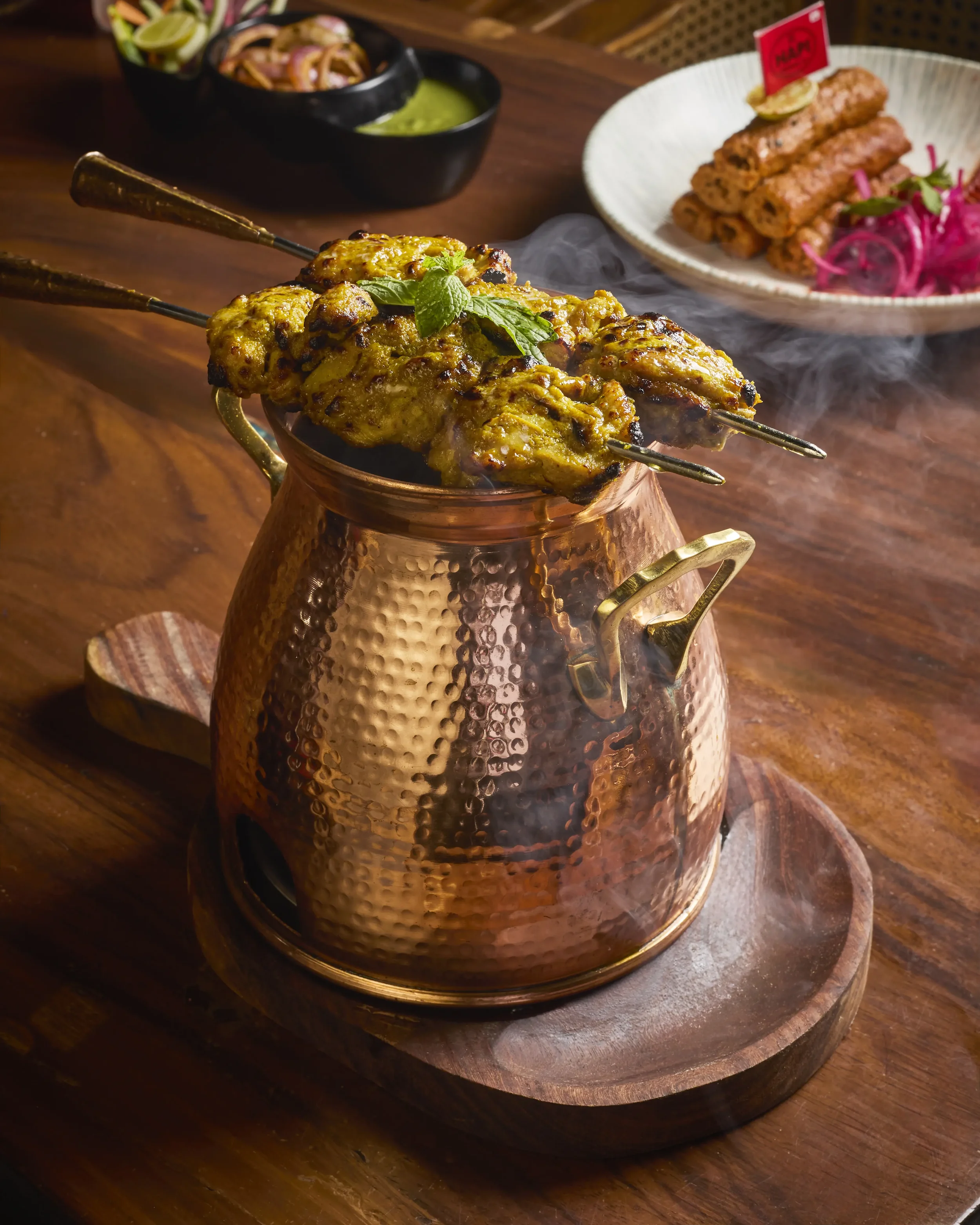 Close-up of skewered grilled chicken pieces garnished with mint leaves in a copper serving vessel on a wooden tray, with side dishes including fried items and salad in the background.