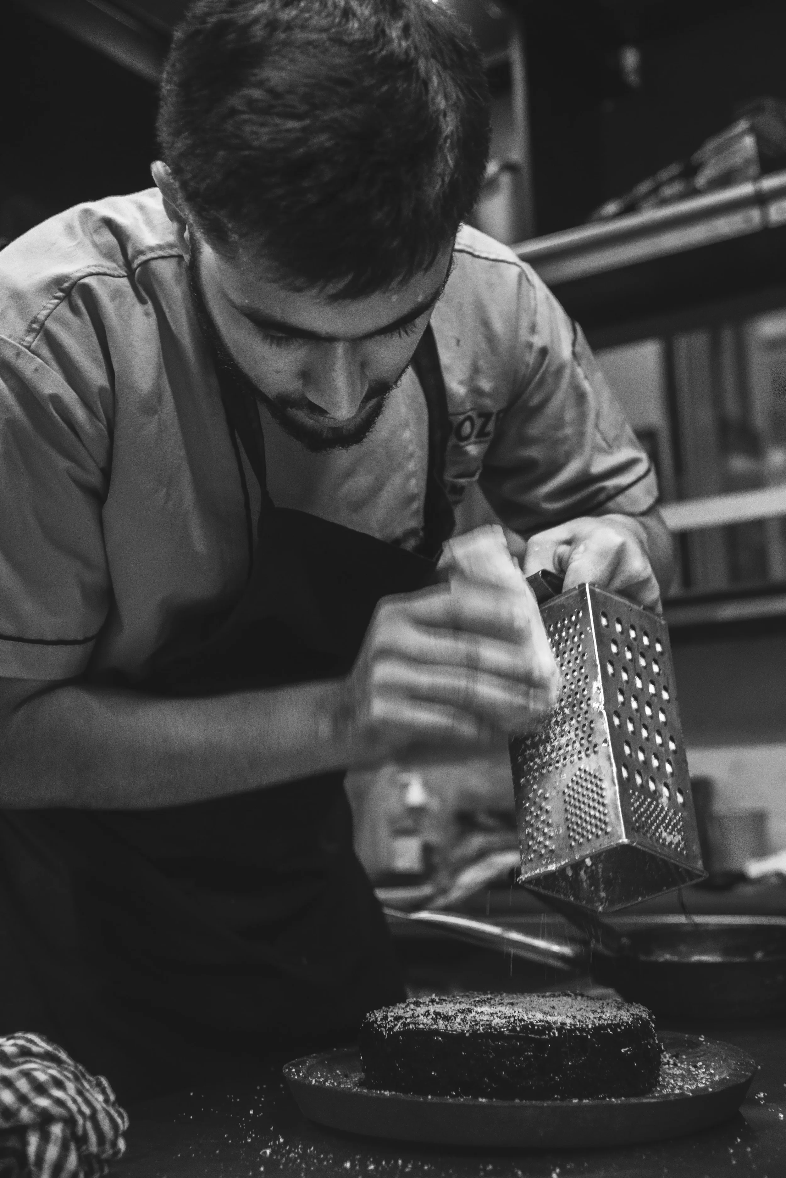 A chef grates cheese over a blackened steak on a plate in a kitchen, in black-and-white.
