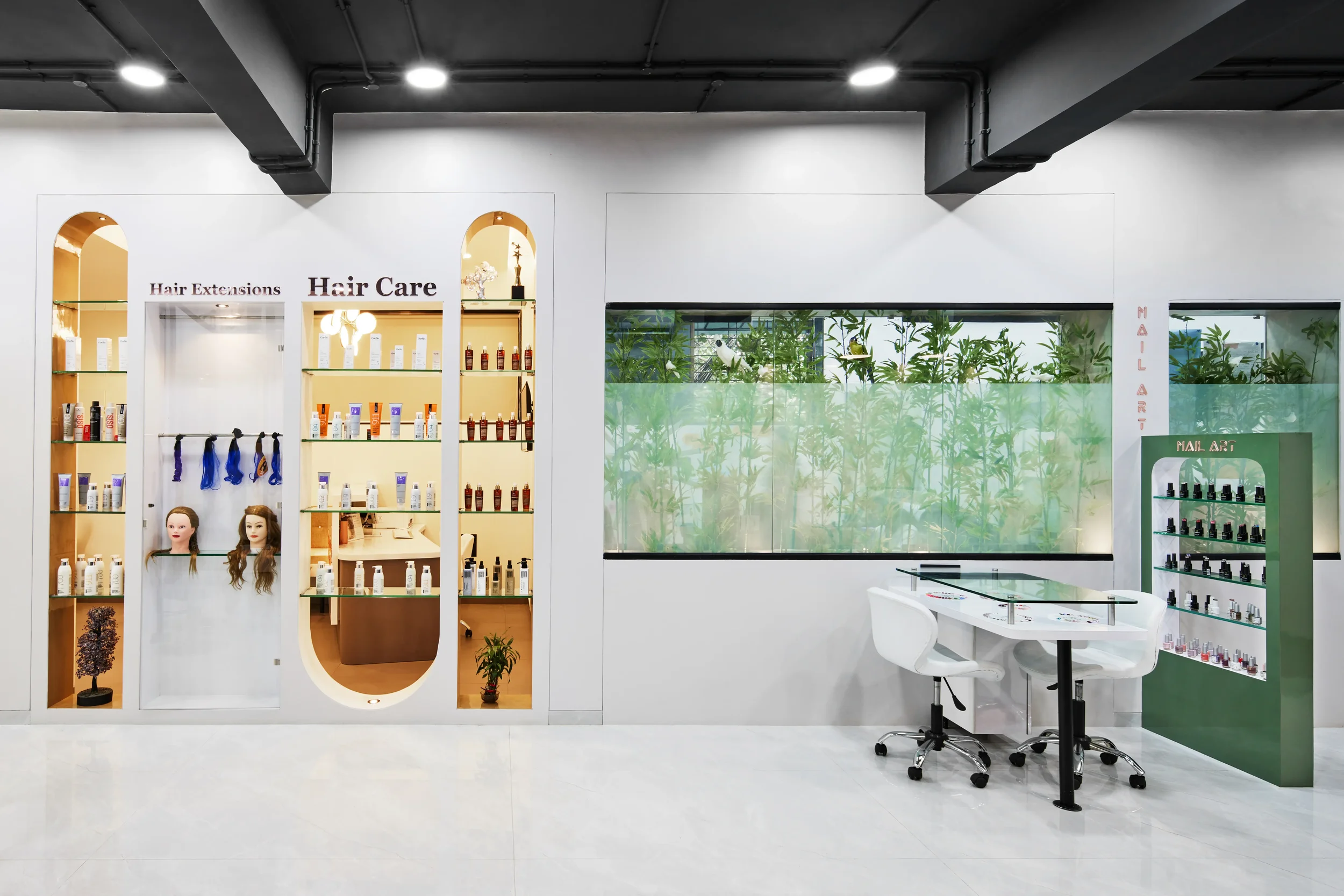 Interior of a beauty salon with shelves of hair care products, mannequin heads with hair extensions, a glass table, and a large window with a view of green plants outside.