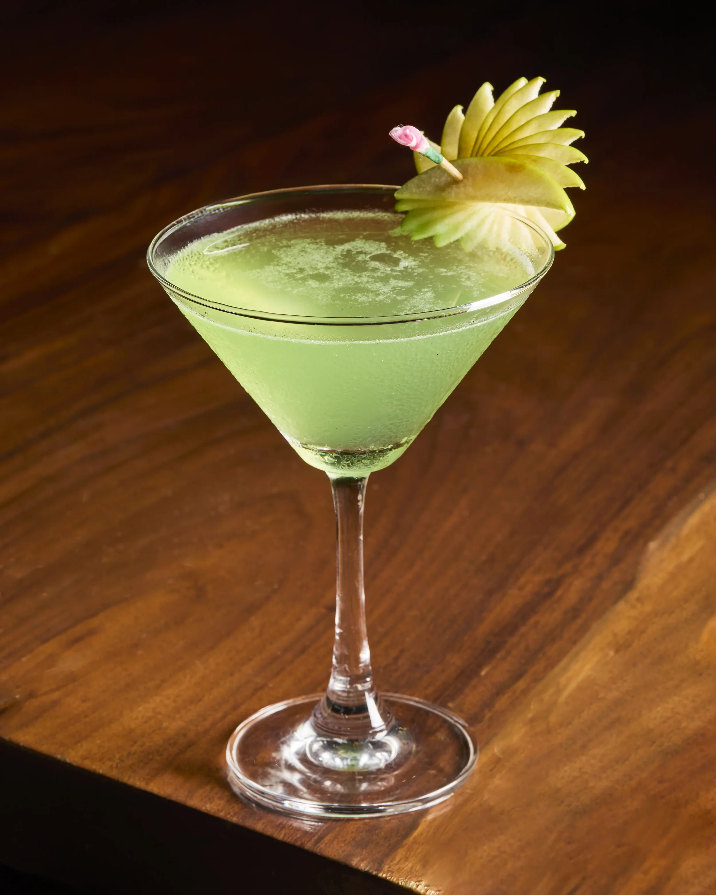 A green cocktail in a martini glass garnished with a carved apple shaped like a flower and a toothpick with a pink decorative item, set on a wooden surface.