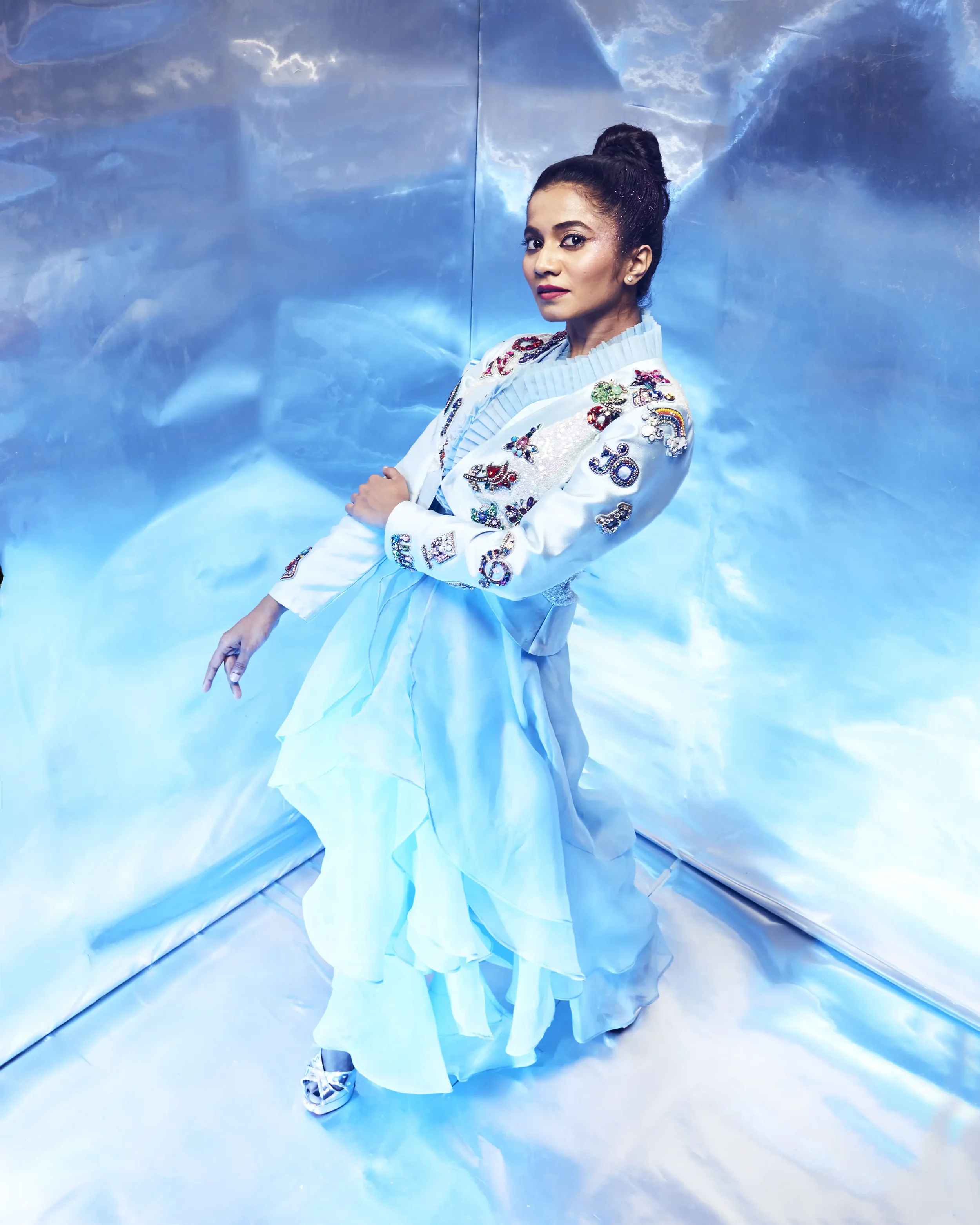 A woman dressed in a light blue, embellished outfit with embroidered patches, standing against a reflective, blue-toned backdrop, posing with arms crossed and looking at the camera.