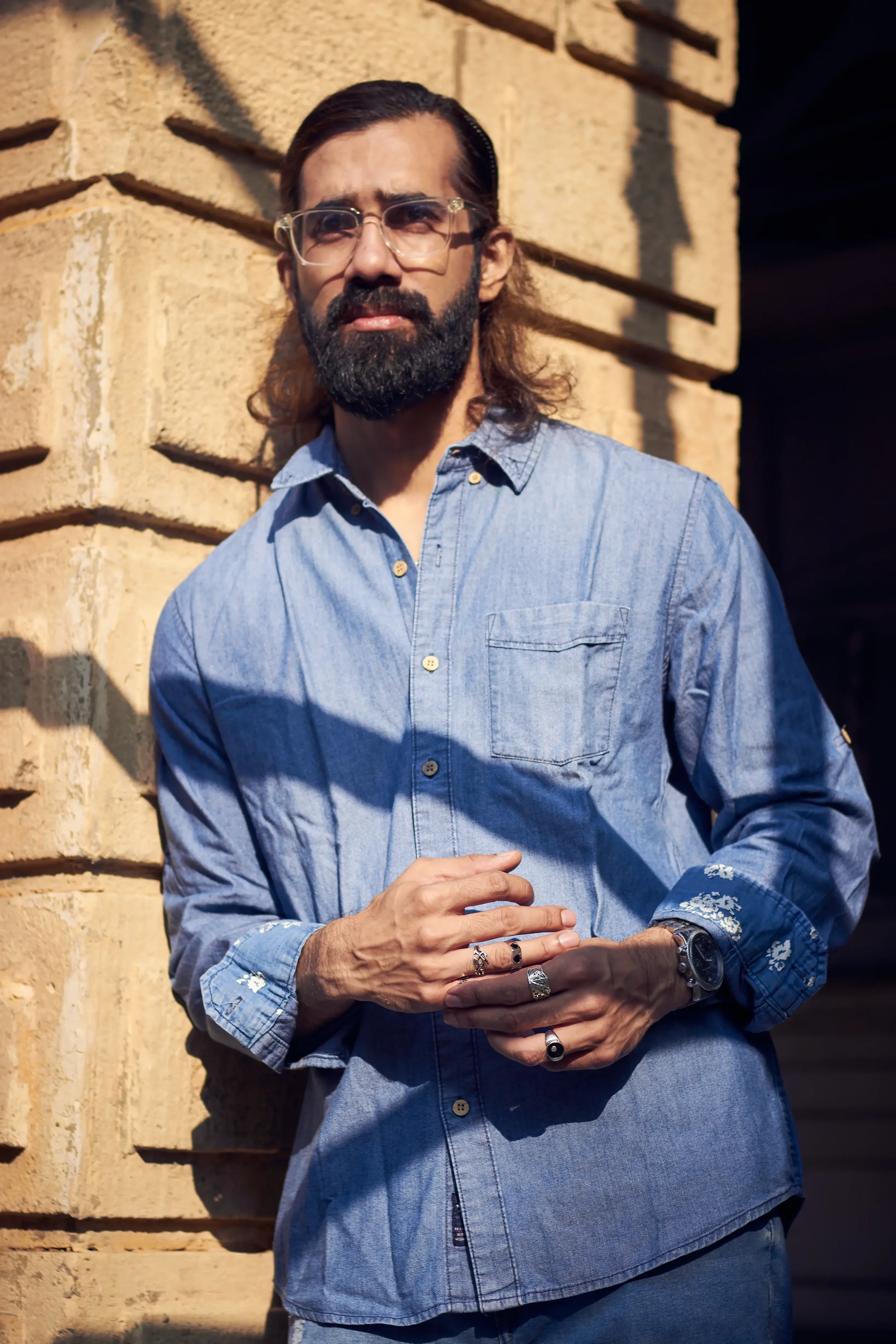 A man with glasses, a beard, and long hair, wearing a blue denim shirt and multiple rings, standing against a brick wall in sunlight.
