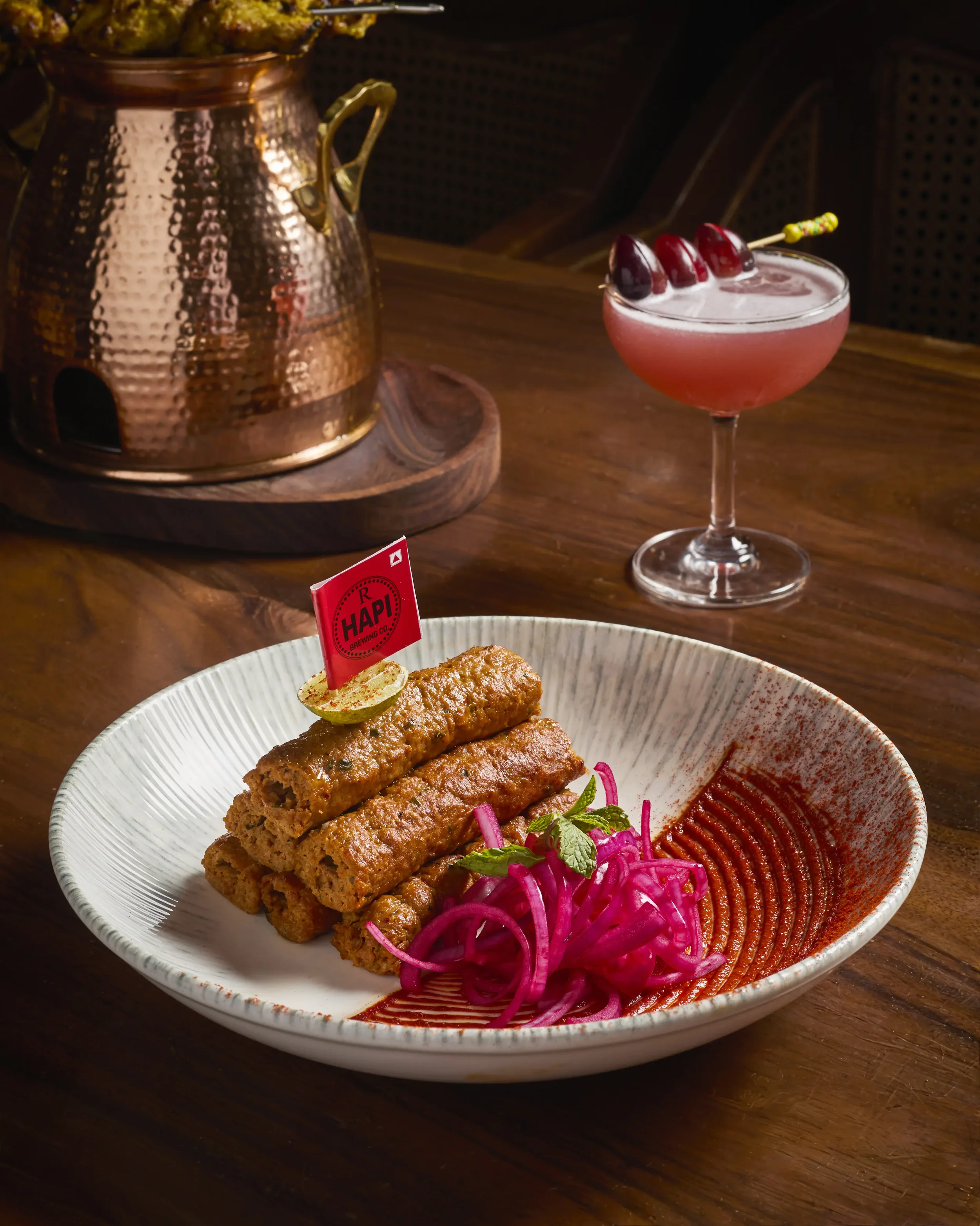 A white bowl filled with mutton seekh kebab, pink pickled onions, colorful herbs, and two pieces of fried food, possibly fish or plantains, with a spoonful of sauce on the side, placed on a dark wooden table.