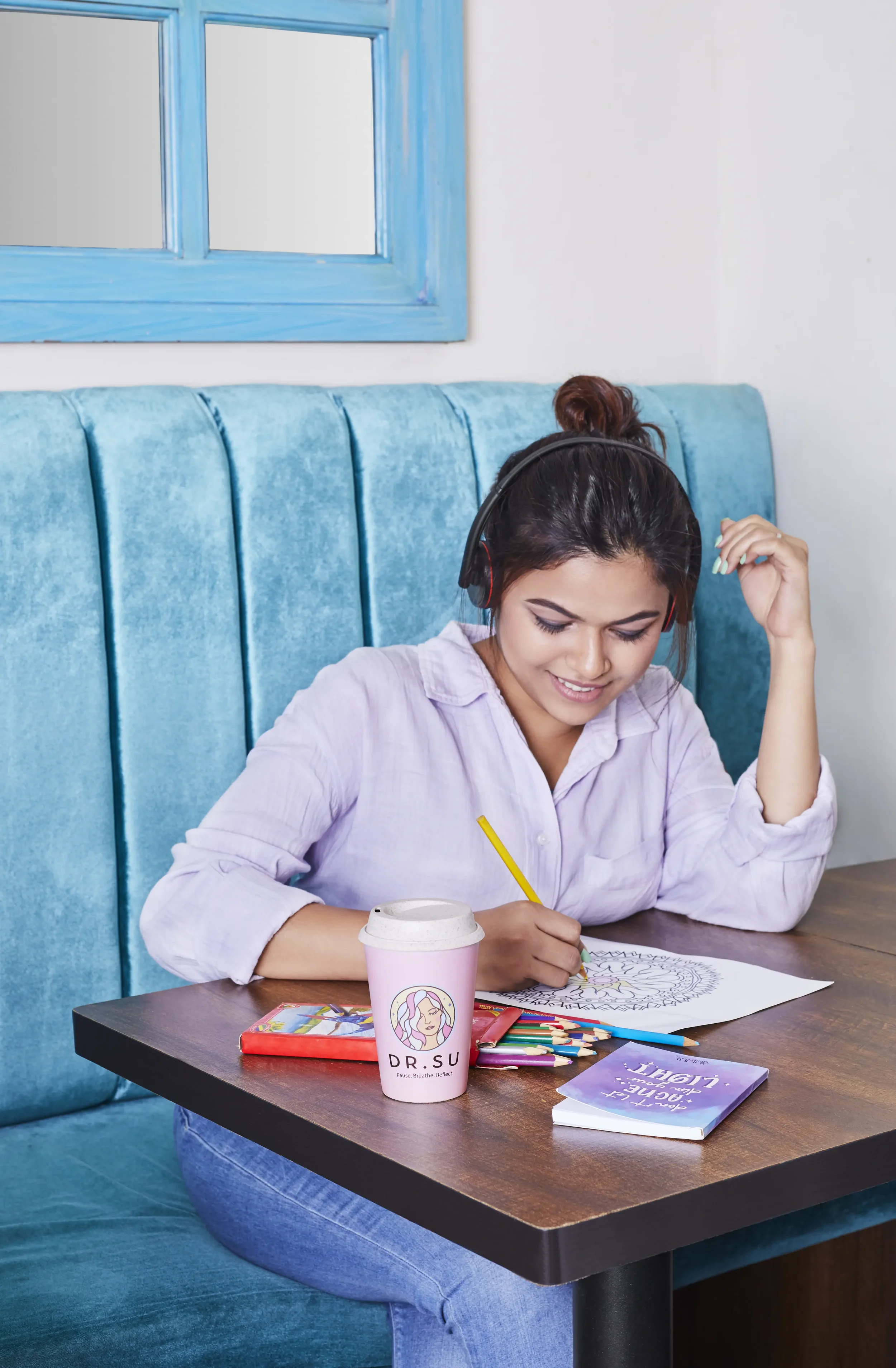A young woman sitting at a table in a cafe, coloring a mandala design on paper, wearing a lavender shirt and black headphones, with a pink coffee cup labeled 'DR. SU' and colored pencils on the table.
