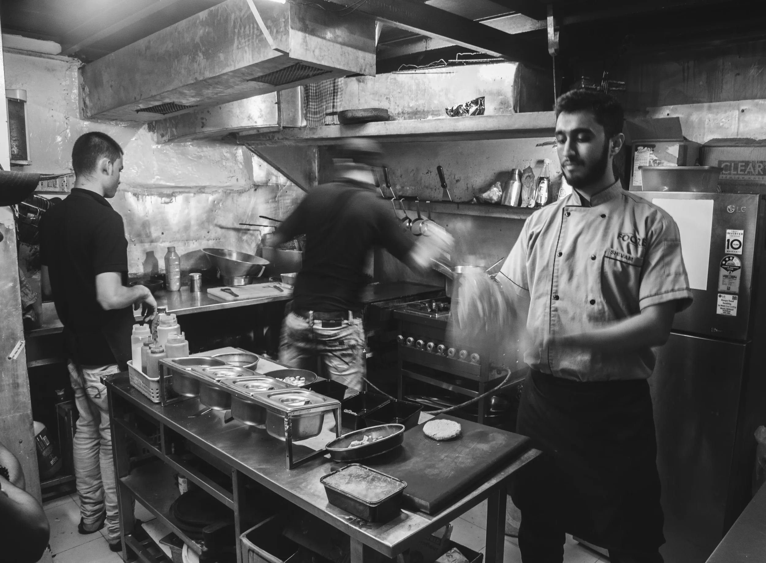 Three men working in a restaurant kitchen, one wearing an apron and chef's coat, another stirring a pan on the stove, and the third preparing ingredients at a counter, all in black and white.