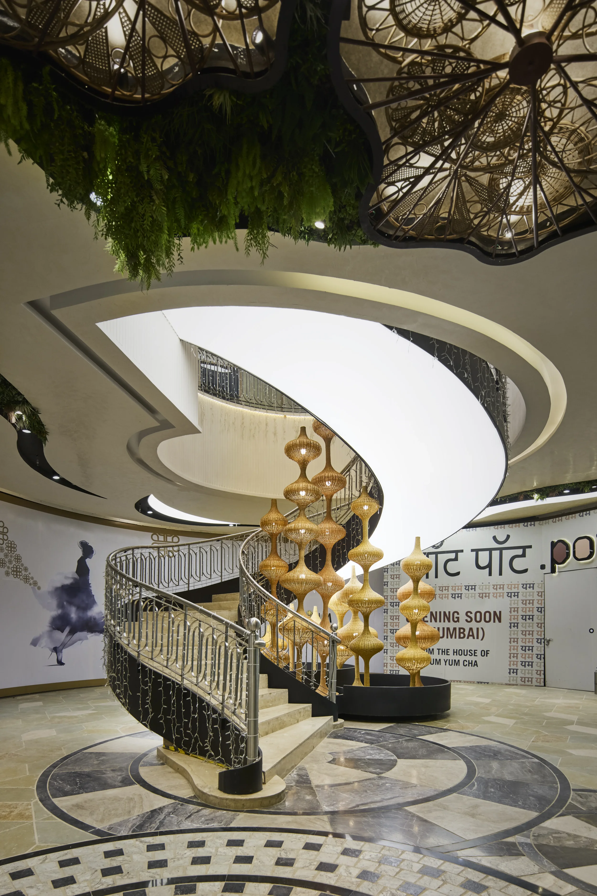 Interior view of a modern mall with a spiral staircase. The ceiling features large woven chandeliers and a prominent curved white ceiling panel. There are green plants hanging from the ceiling and a promotional poster in the background.