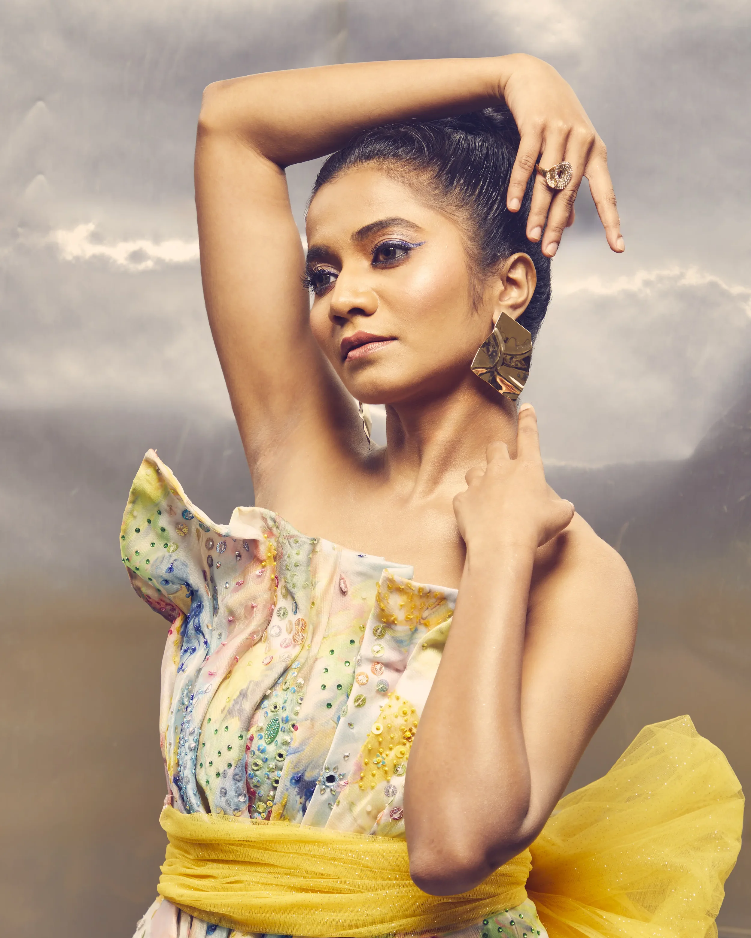 A woman with dark hair styled in a bun, wearing a colorful off-shoulder dress with intricate embellishments, large gold earrings, and a yellow belt or sash, standing against a cloudy sky background.