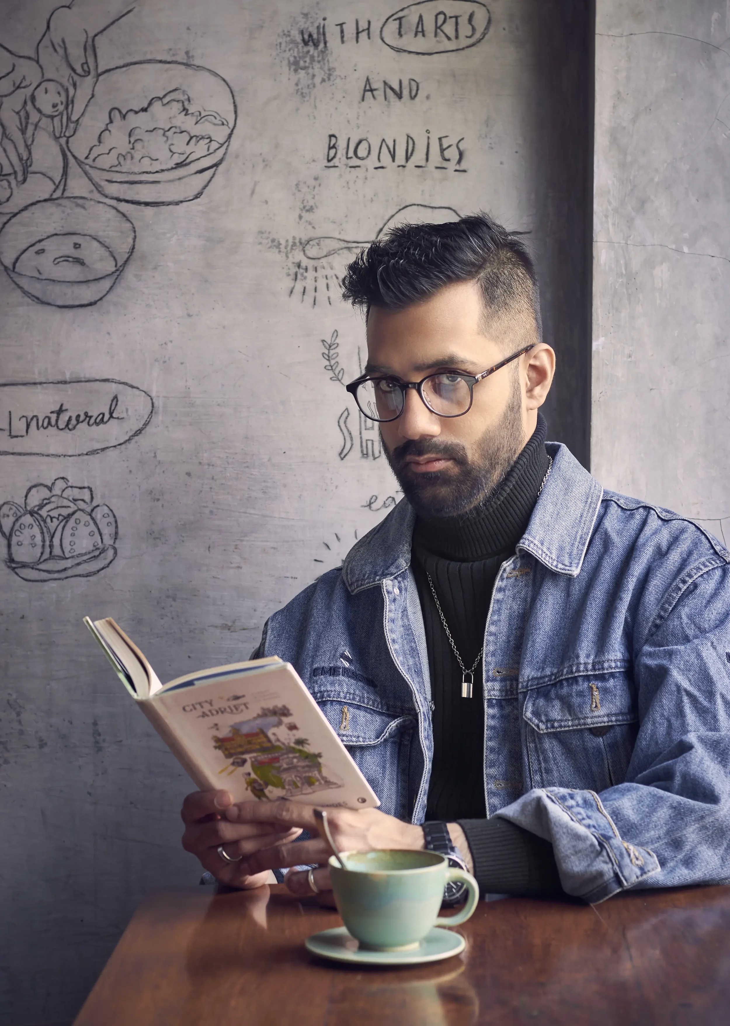 A man with glasses and a denim jacket sitting at a wooden table, reading a colorful travel guidebook with a cup of tea or coffee in front of him. Behind him is a chalkboard wall with drawings and text about food with tart and blondies, and the word '