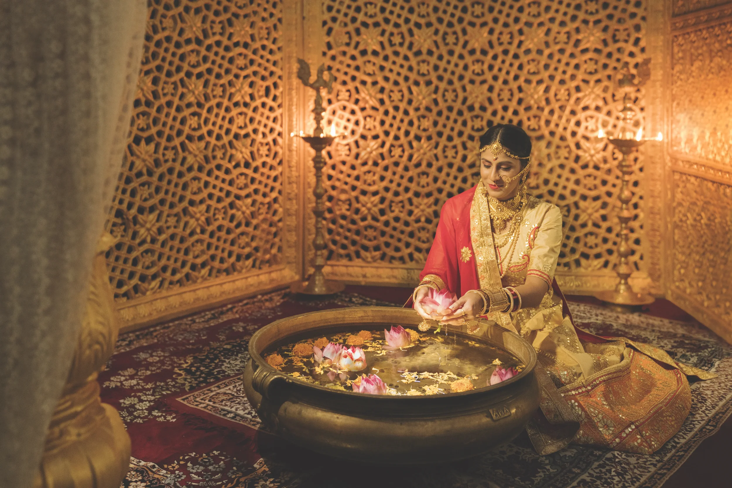 A woman dressed in traditional Indian attire, wearing gold jewelry, sitting on the floor of a richly decorated room, holding a lotus flower over a water basin filled with floating flowers, surrounded by ornate wooden screens and lit by candlelight.