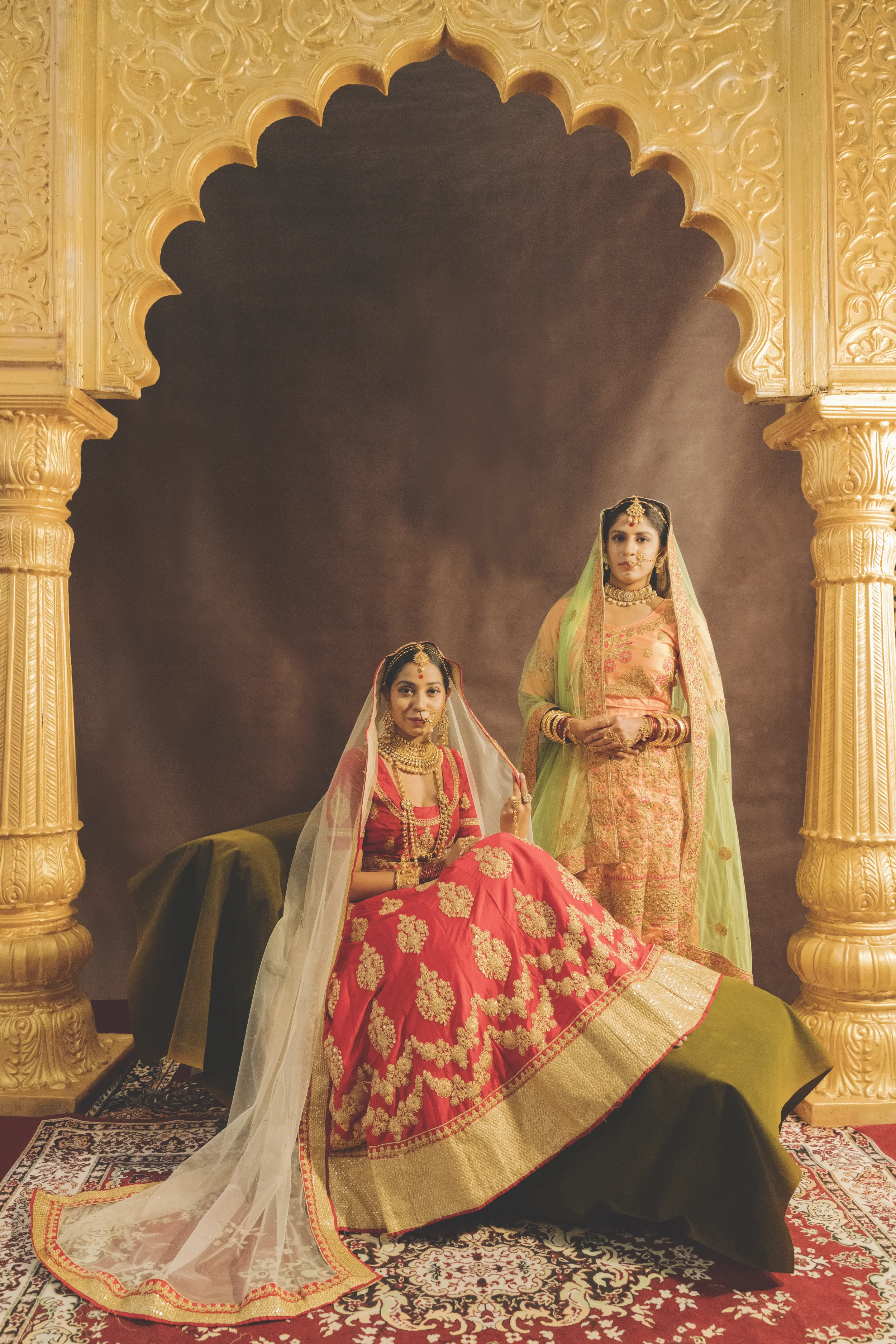 Two women dressed in traditional Indian bridal attire, one seated and one standing, in an ornately decorated setting with golden arches.