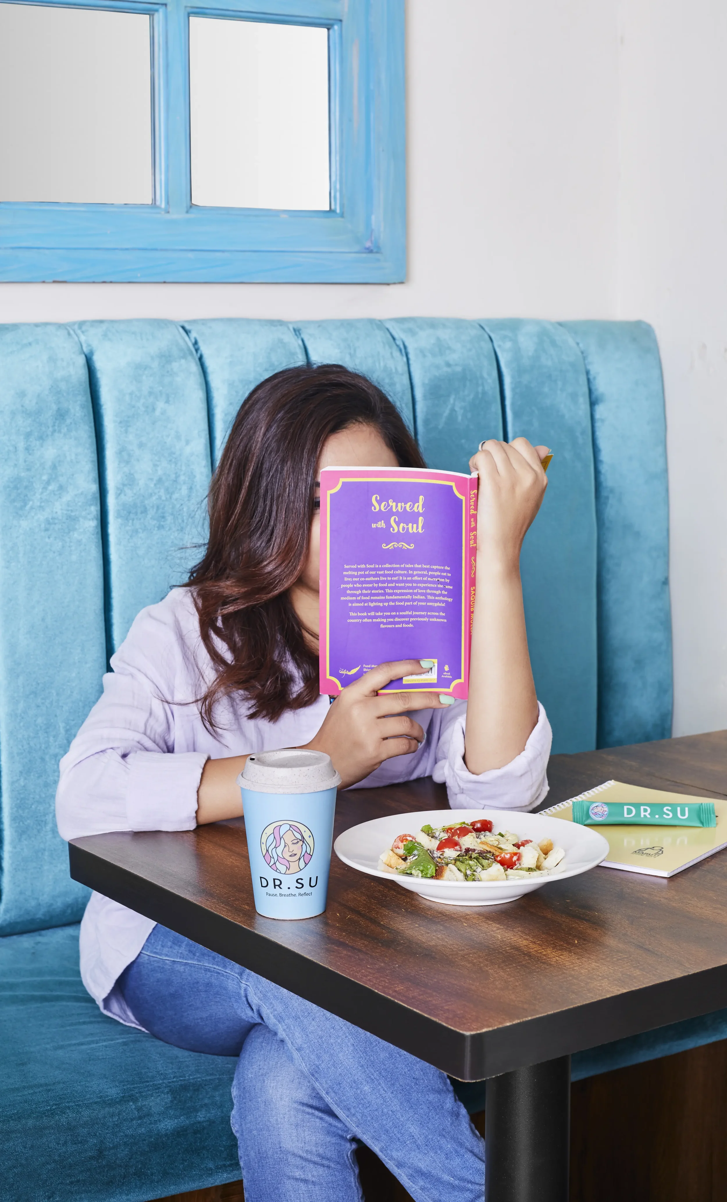 A woman sitting in a blue booth at a restaurant, holding a purple and pink book covering her face. On the table are a salad, a blue coffee cup labeled 'DR. SU', a green and white tube labeled 'DR. SU', and a notebook.