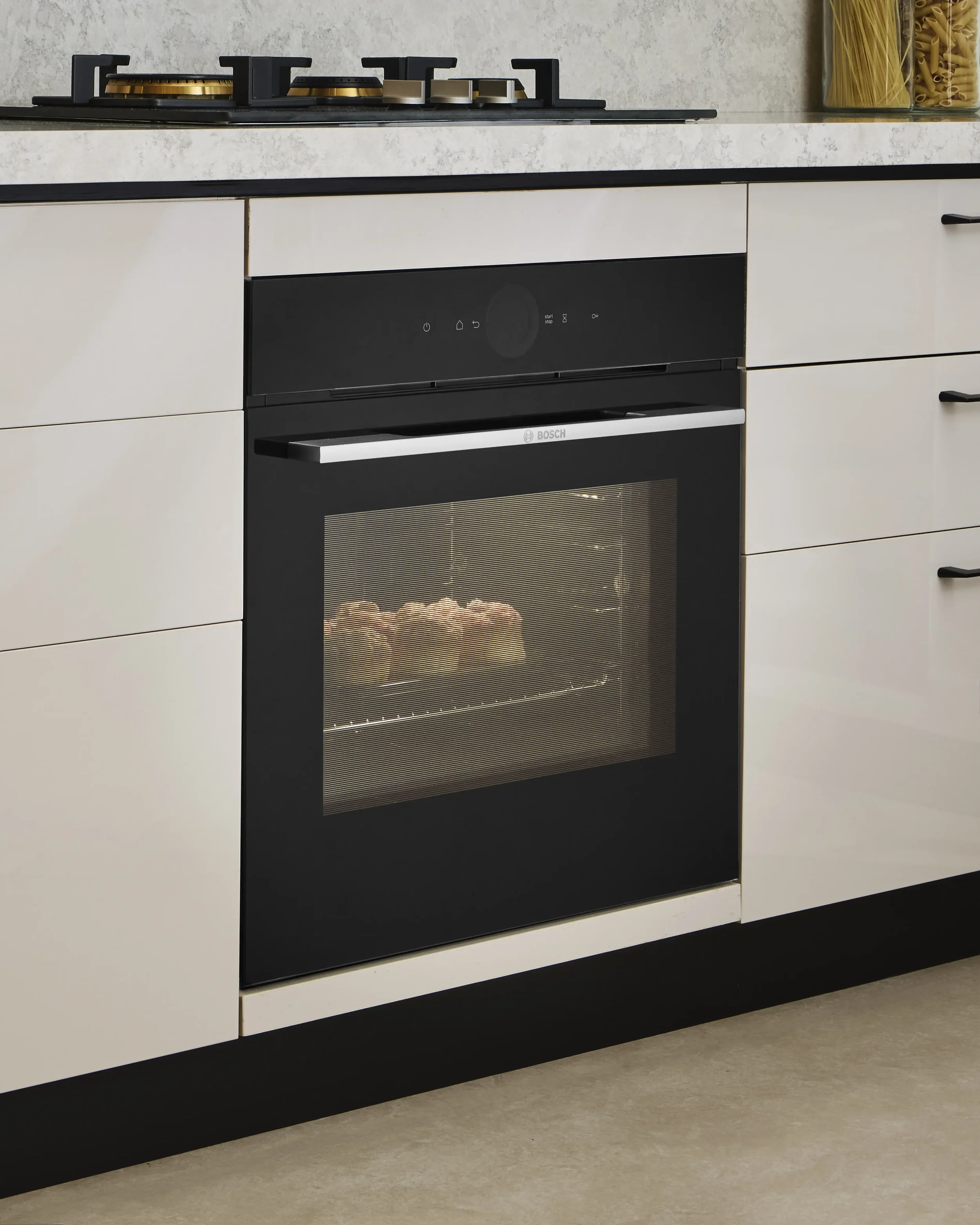 A modern kitchen with a black and white oven, stove, and beige cabinets. The oven has a glass door with items visible inside, and the stove has four burners with gold-colored accents. There is a counter on top with a jar of uncooked pasta and a jar o