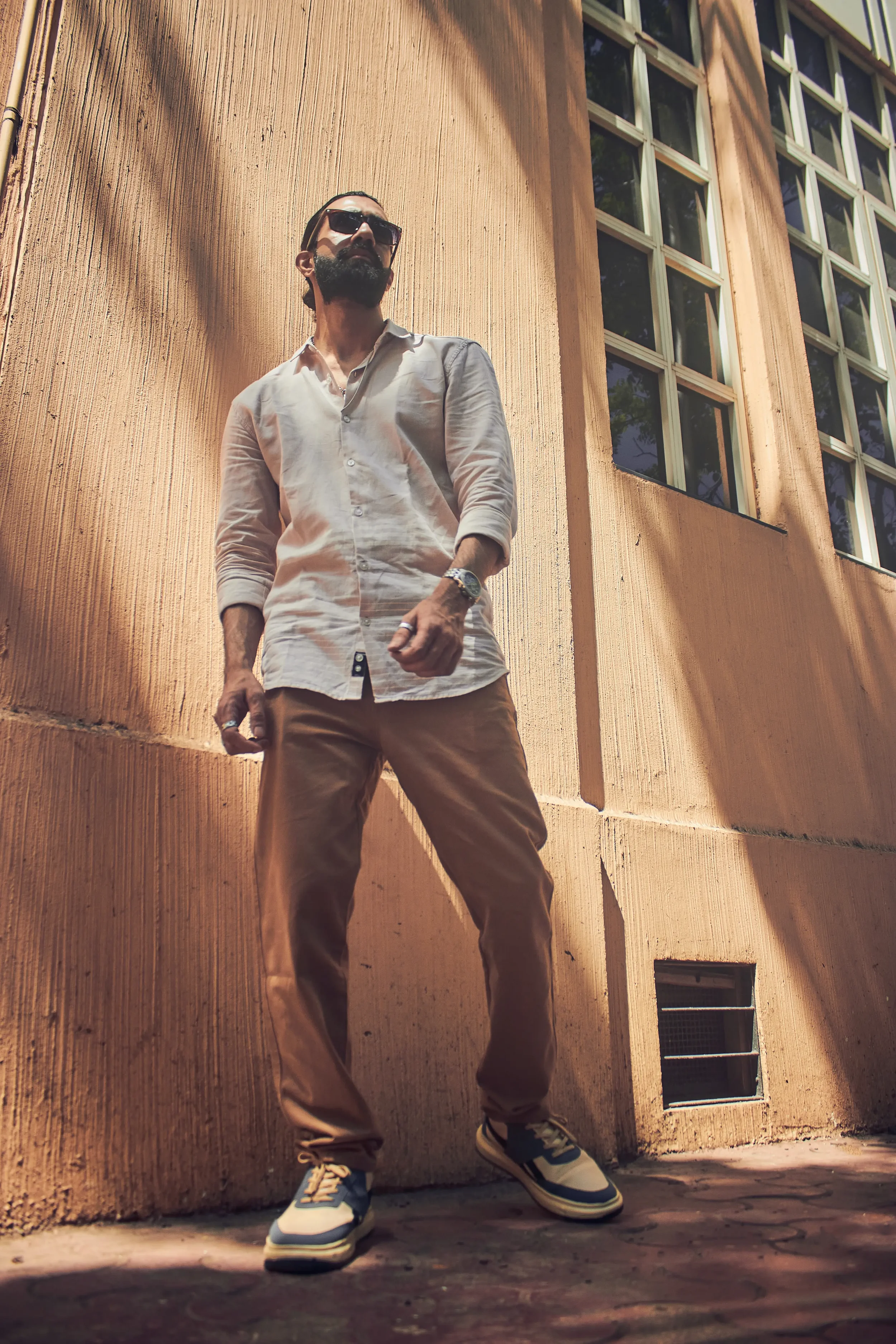 A man stands outdoors against a textured peach-colored wall with tall windows, wearing sunglasses, a light-colored shirt, tan pants, and sneakers.