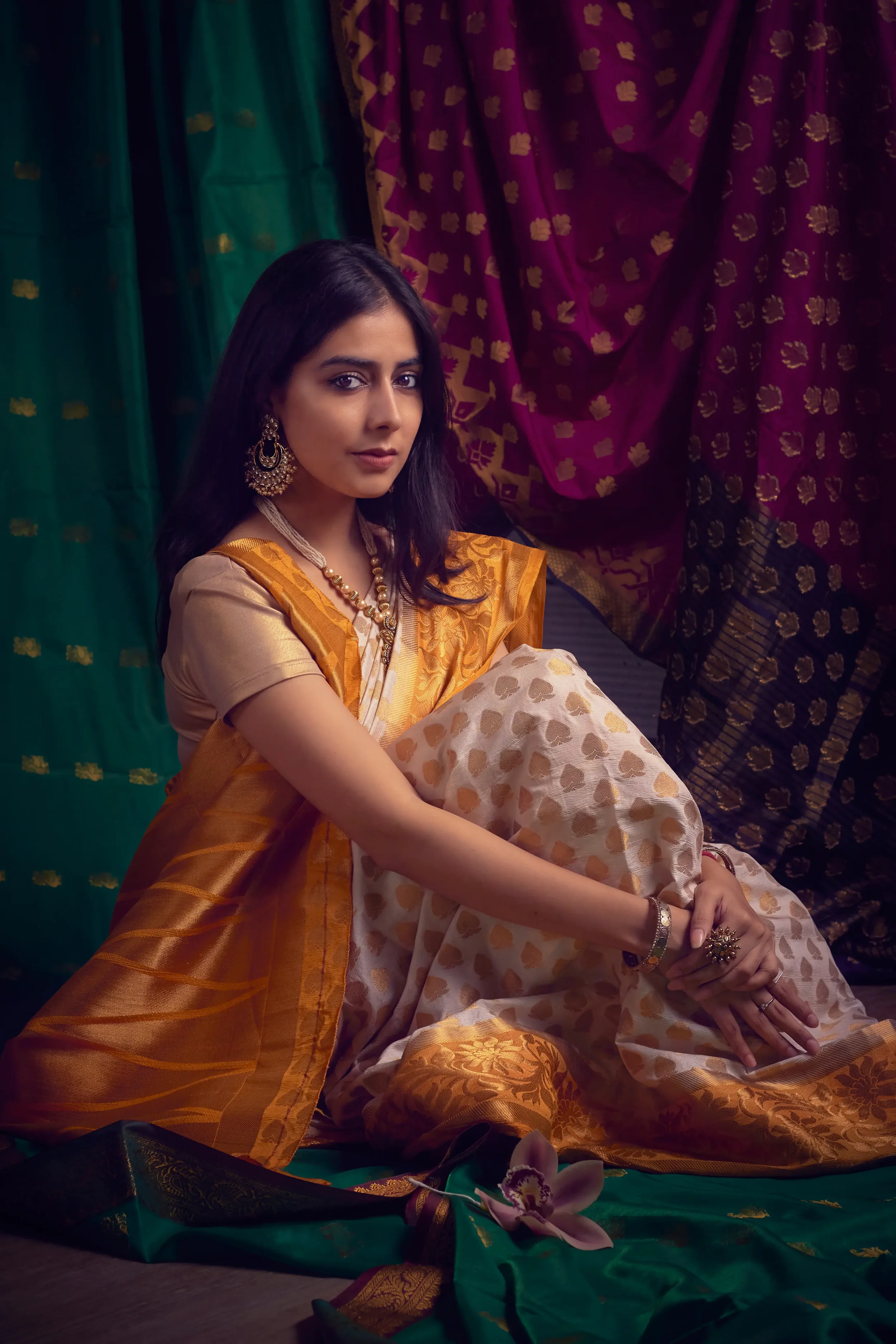 A woman dressed in traditional Indian attire, sitting on the floor with colorful, ornate fabric and flowers around her, against a backdrop of patterned curtains.
