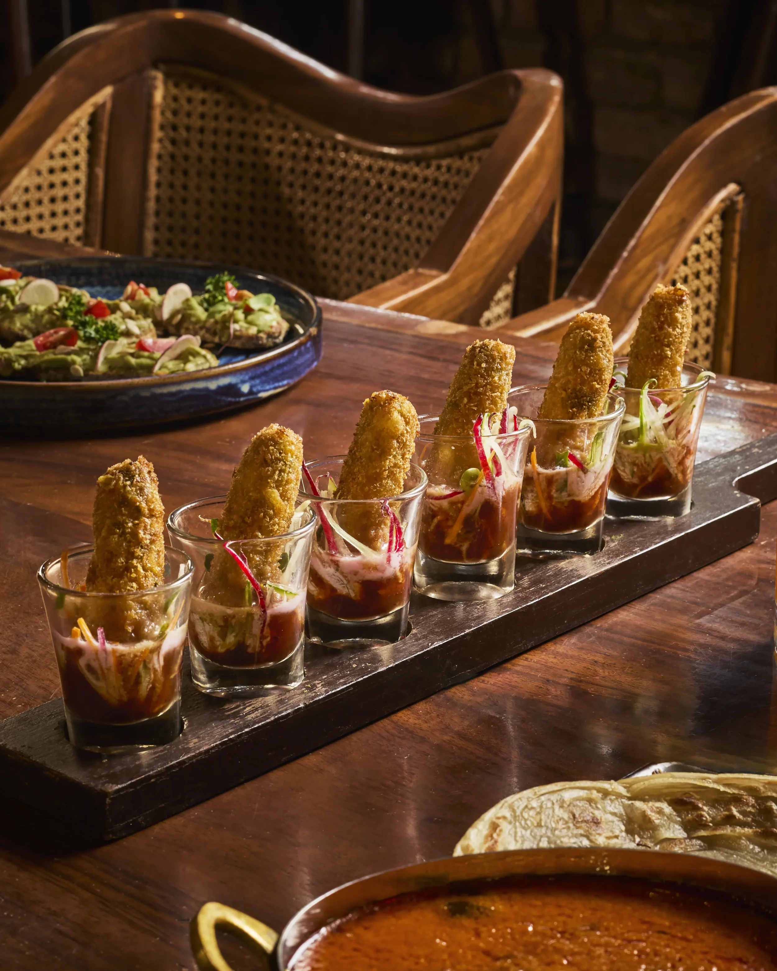 A wooden table with a black serving platter of sliced avocado topped with tomatoes, onion, and herbs. In front, four small glass cups contain crispy breaded appetizers garnished with microgreens. In the background, a bowl of red sauce or salsa and a 