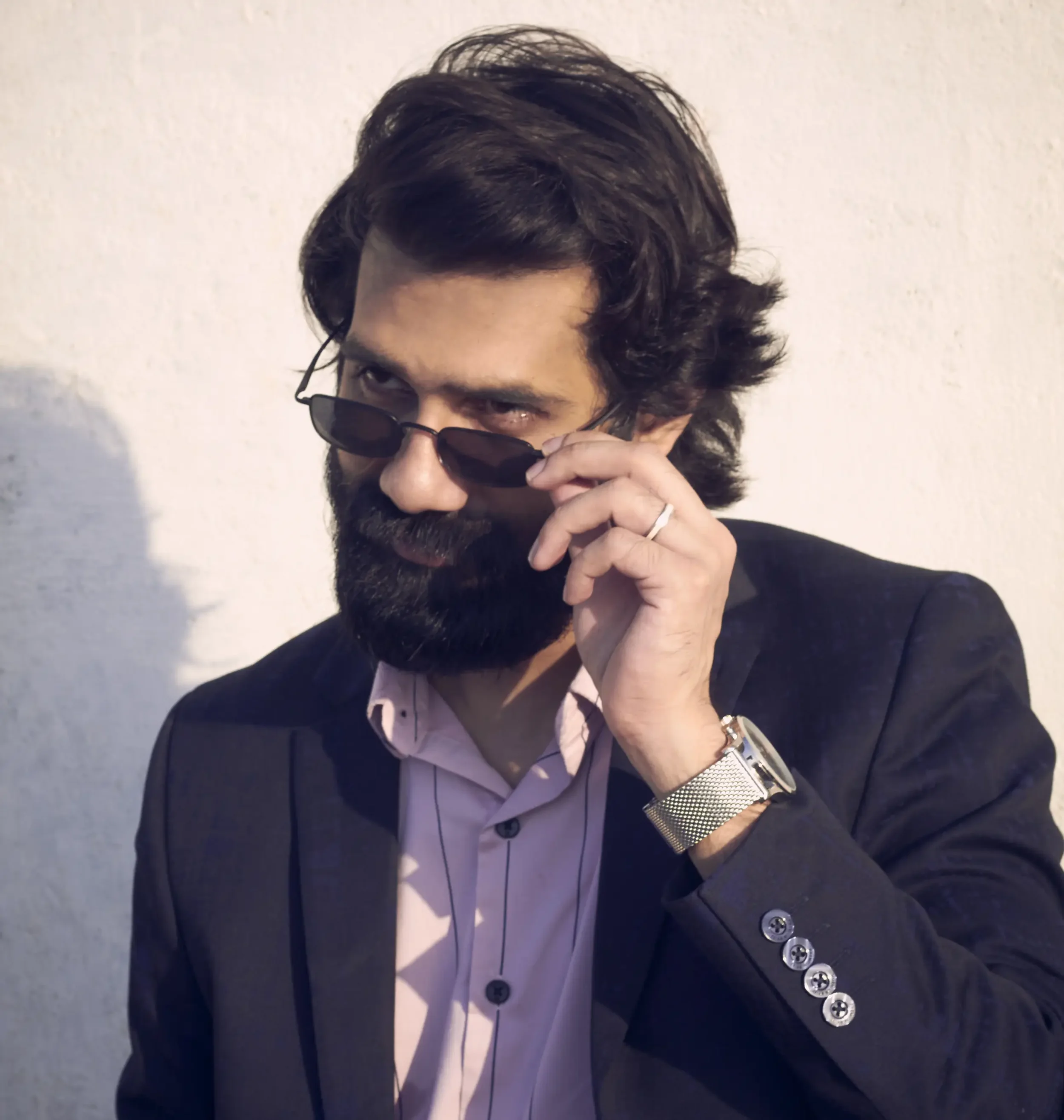 A man with dark hair and a beard wearing a black blazer, pink shirt, sunglasses, and a silver watch, looking over the sunglasses at the camera.