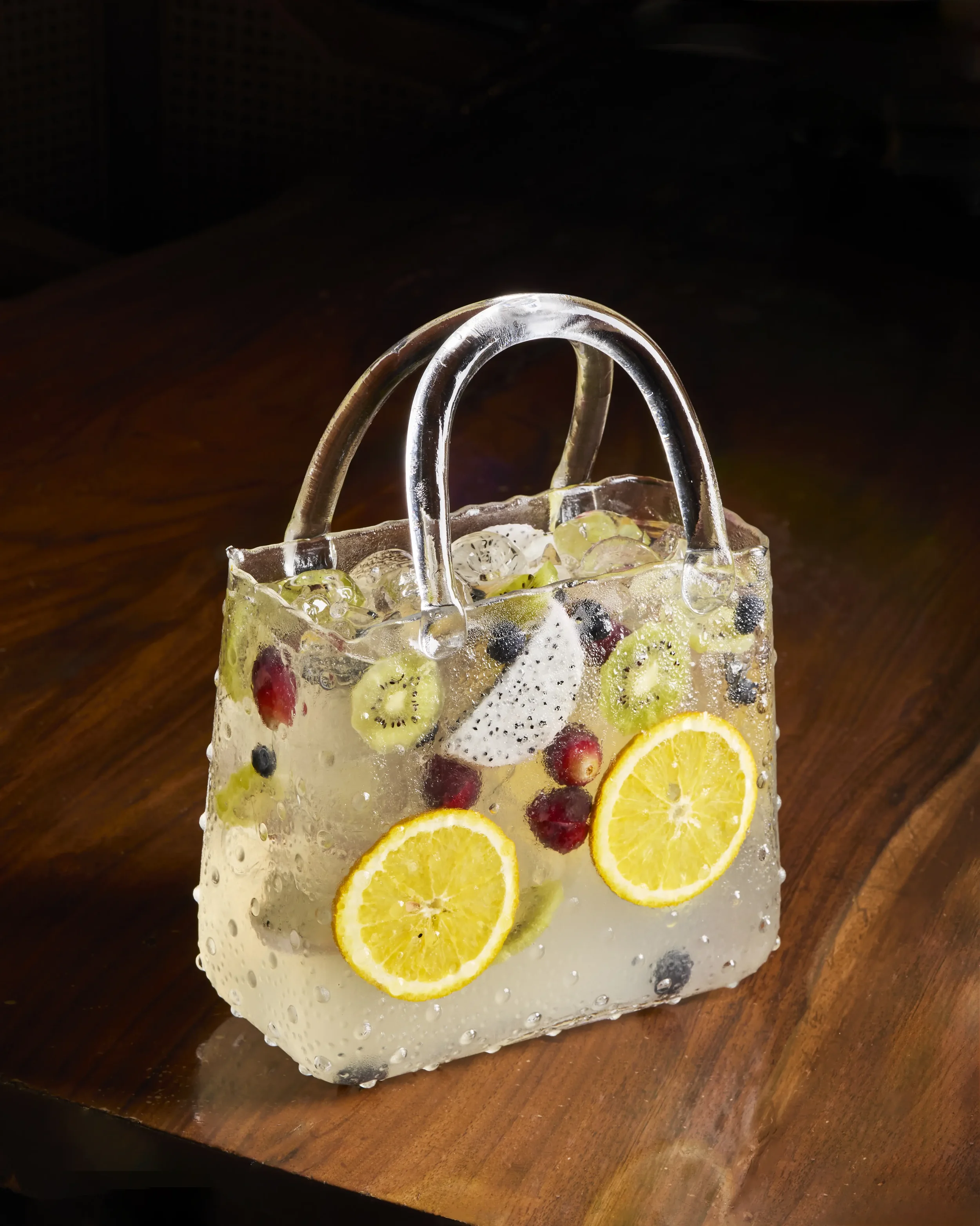 A clear glass or acrylic container filled with ice, lemon slices, orange slices, and assorted berries, with two silver handles at the top, sitting on a wooden surface.