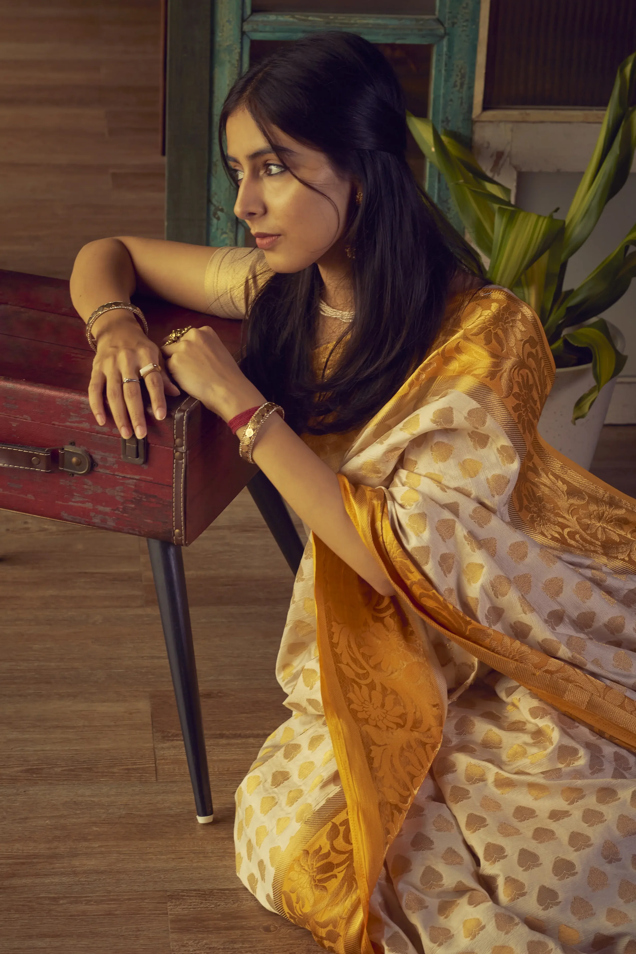 Woman wearing a golden saree with floral and heart patterns, leaning on a wooden table, with a glass of greenery in the background.
