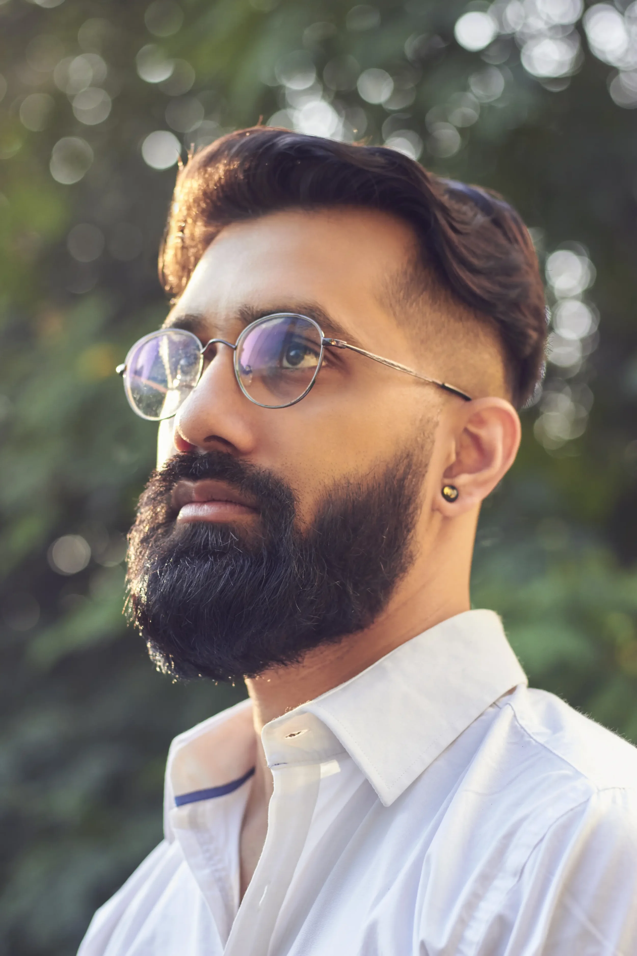 A young man with a beard and glasses looking to the side outdoors with blurred trees in the background.