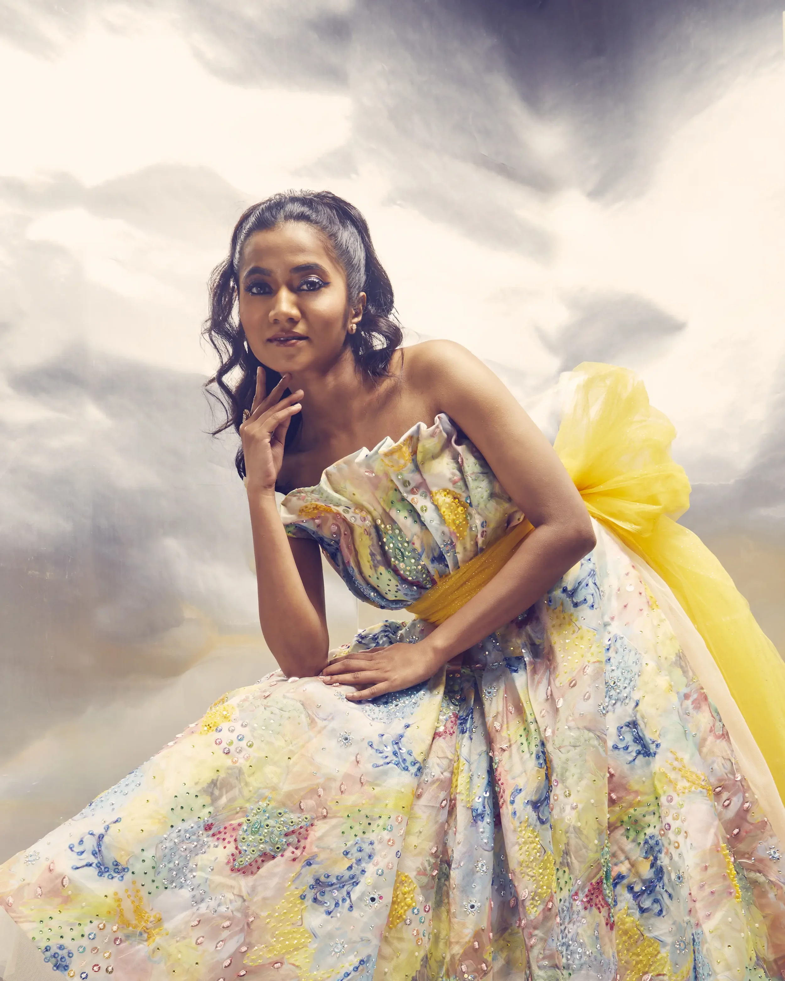 A woman with dark, wavy hair tied back, wearing a colorful, embellished dress with a large yellow bow at the back, sitting against a cloudy sky background.