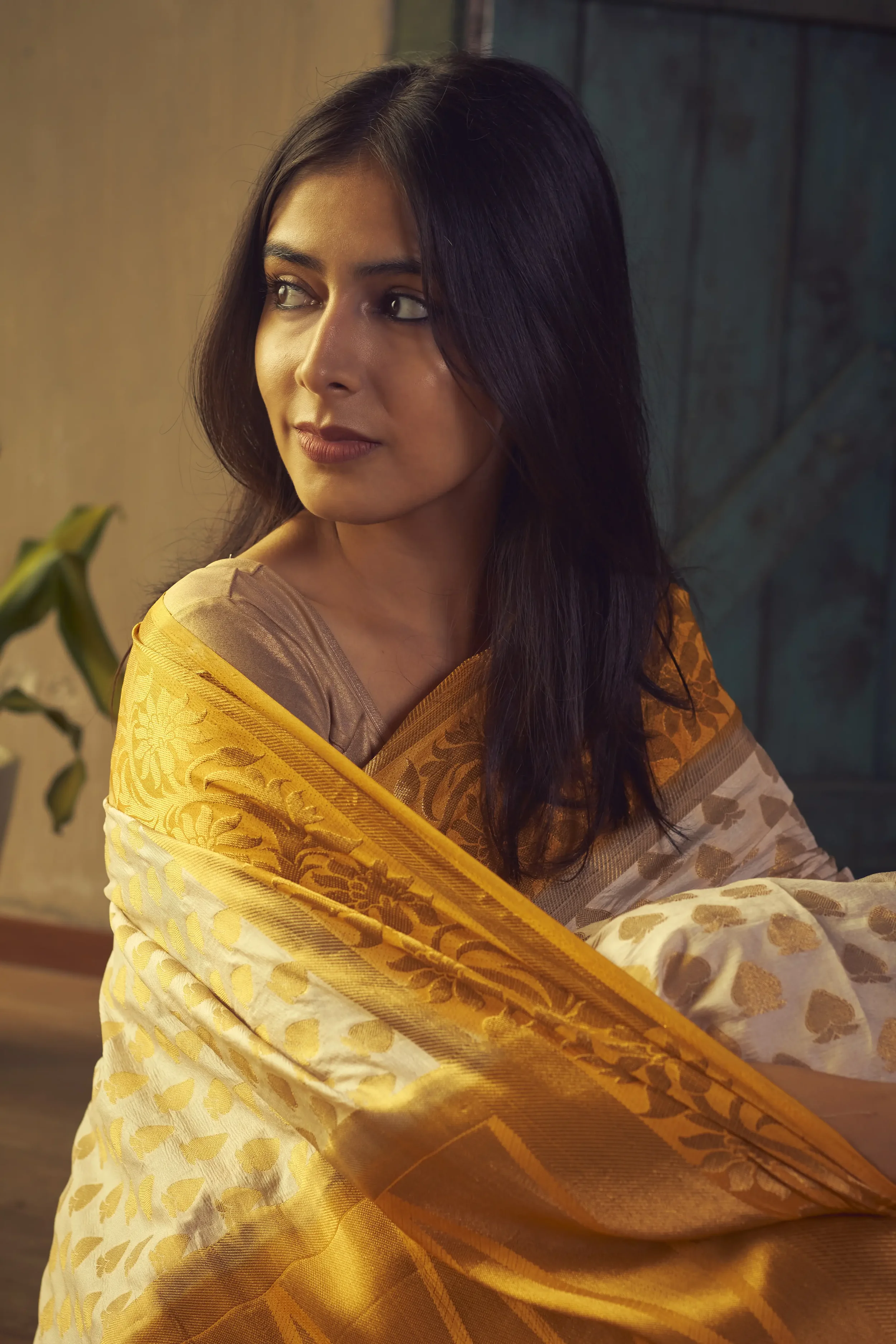 A woman with long dark hair wearing a yellow and cream sari with floral patterns, sitting and looking to her right in a warmly lit room.