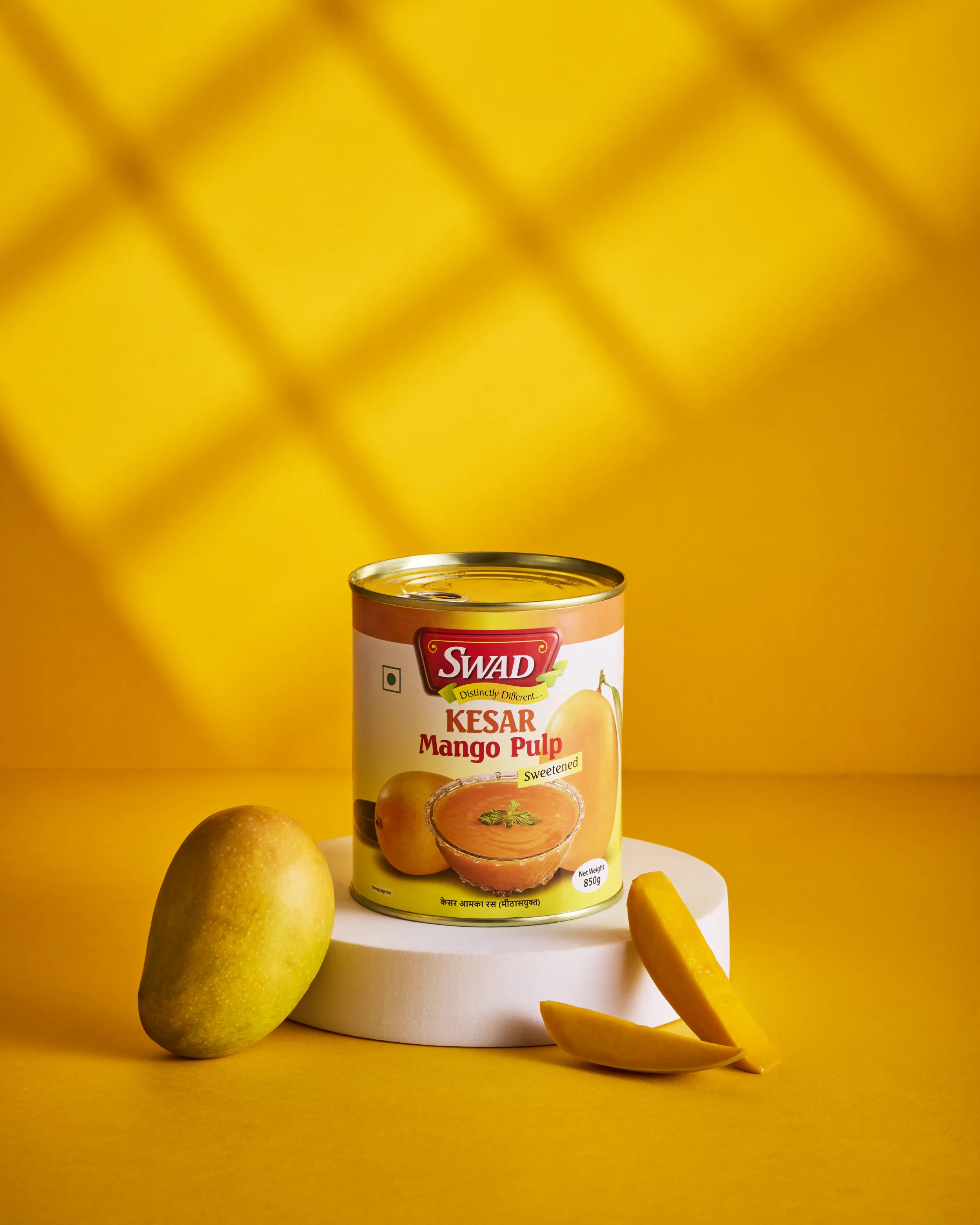 A can of Swad Kesar Mango Pulp on a white cylindrical platform, with two whole mangoes and a mango slice in front, against a yellow background.