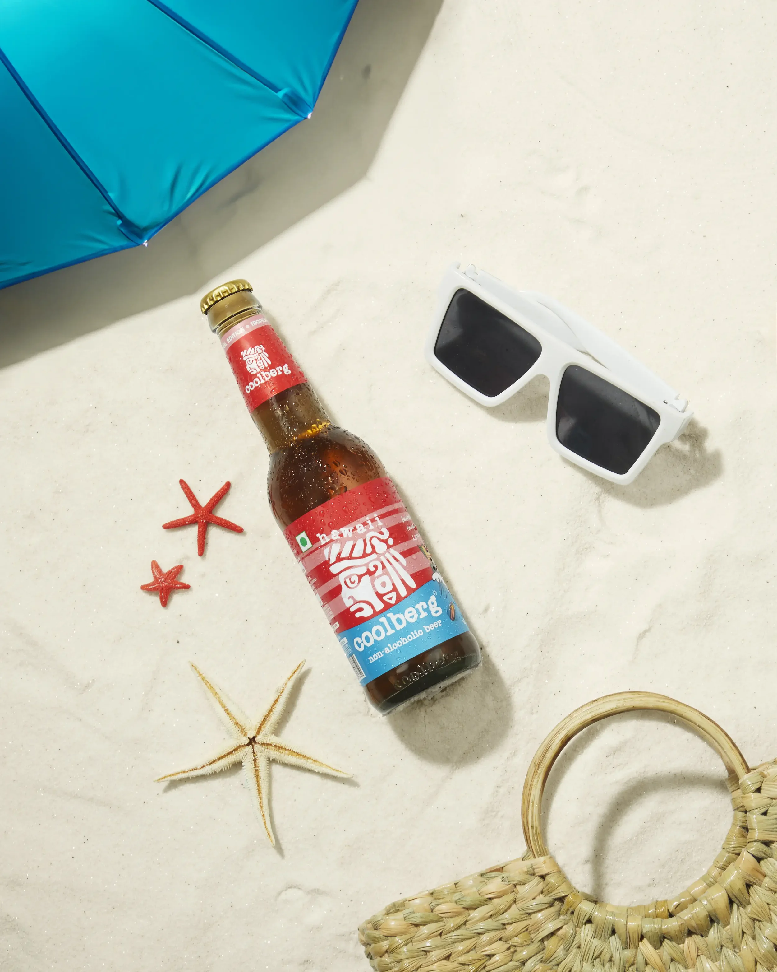 Beach scene with sunglasses, a Coolberg non-alcoholic beer bottle, starfish, seashells, a woven straw bag, and a blue beach umbrella on sand.