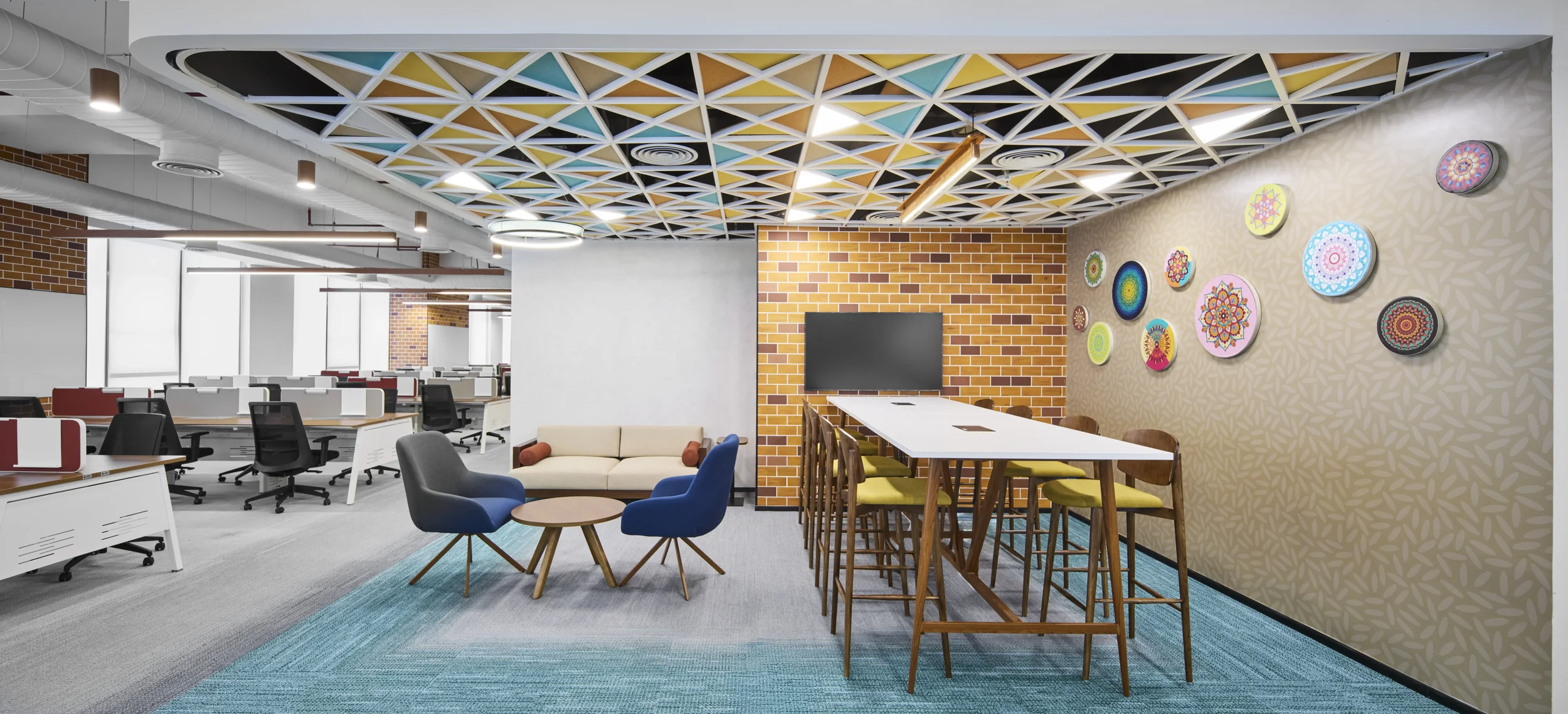 Open office space with desks, chairs, a sitting area with a sofa and chairs, a wall with colorful mandala art, and decorated ceiling panels.