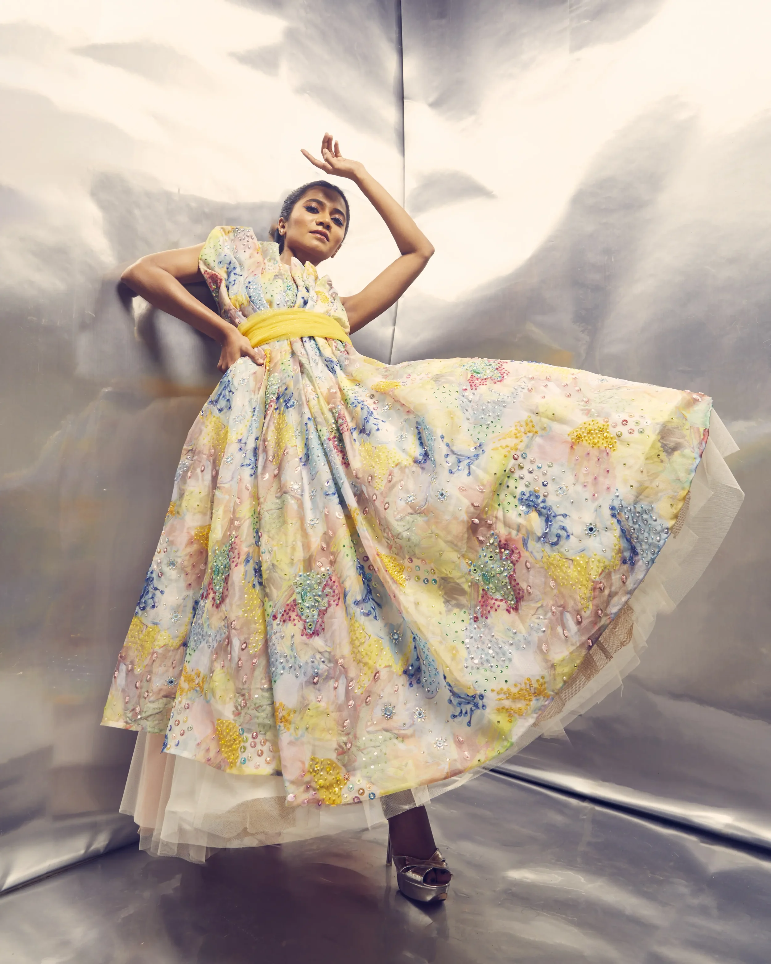 A woman wearing a colorful, embellished gown with a yellow sash, posing with one hand on her hip and the other above her head, in front of a metallic backdrop.