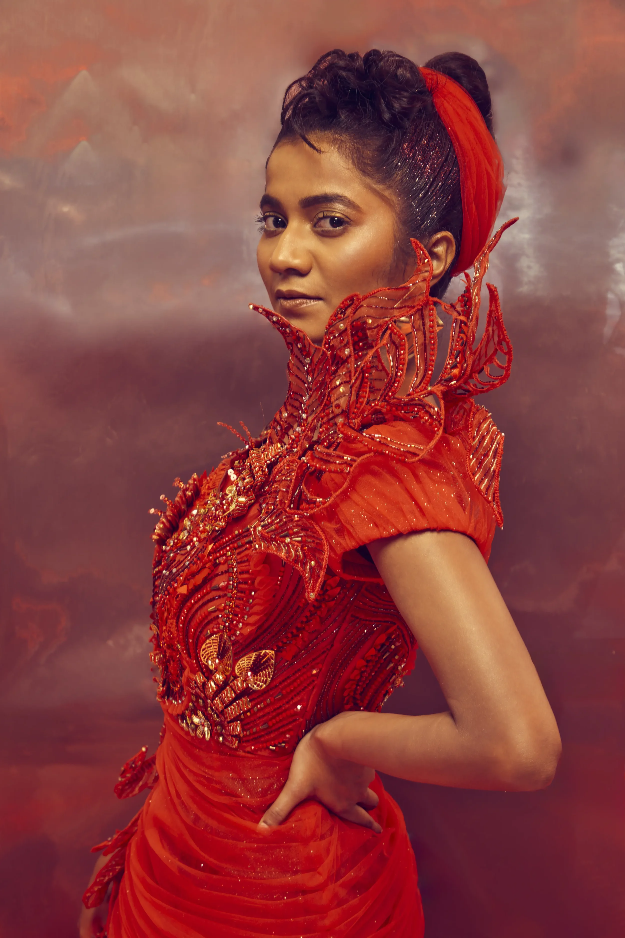 A woman dressed in an elaborate red outfit with intricate beading and fabric details, wearing a red headscarf, standing against a soft pink and brown background.