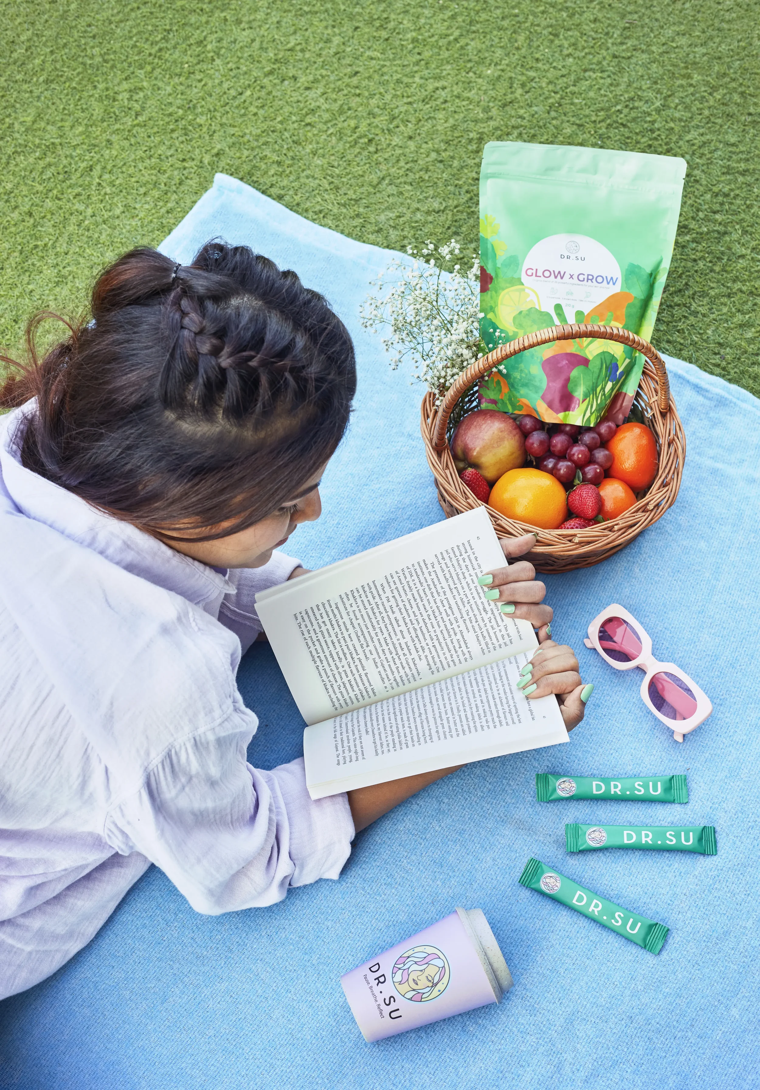 A woman with braided hair reads a book while lying on a blue blanket outdoors. Nearby, there is a basket of fresh fruit, sunglasses, and Dr. Su skincare products. A green packet labeled 'Glow x Grow' is also in the basket.