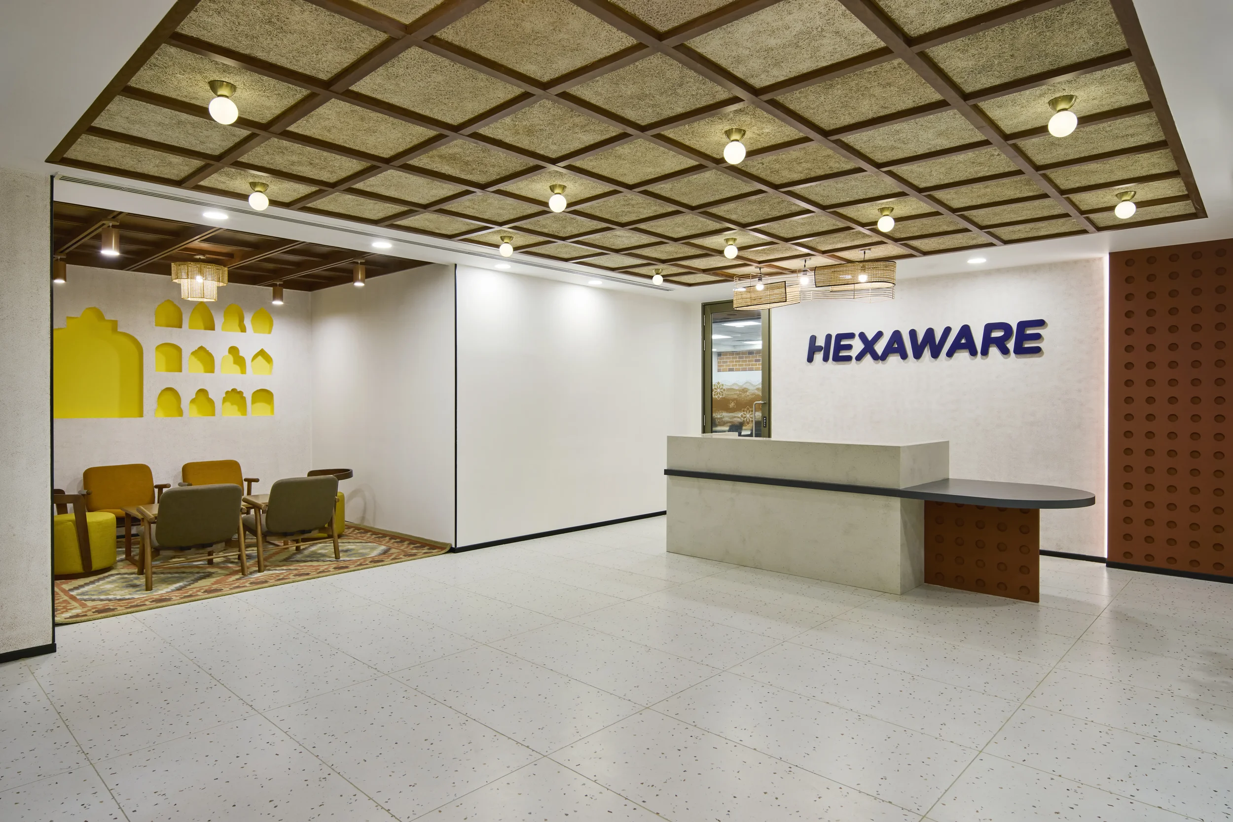 Interior view of a reception area with a white desk, a sign that says 'HEXAWRE', seating with yellow and gray chairs, decorative lighting fixtures, and patterned ceiling and walls.