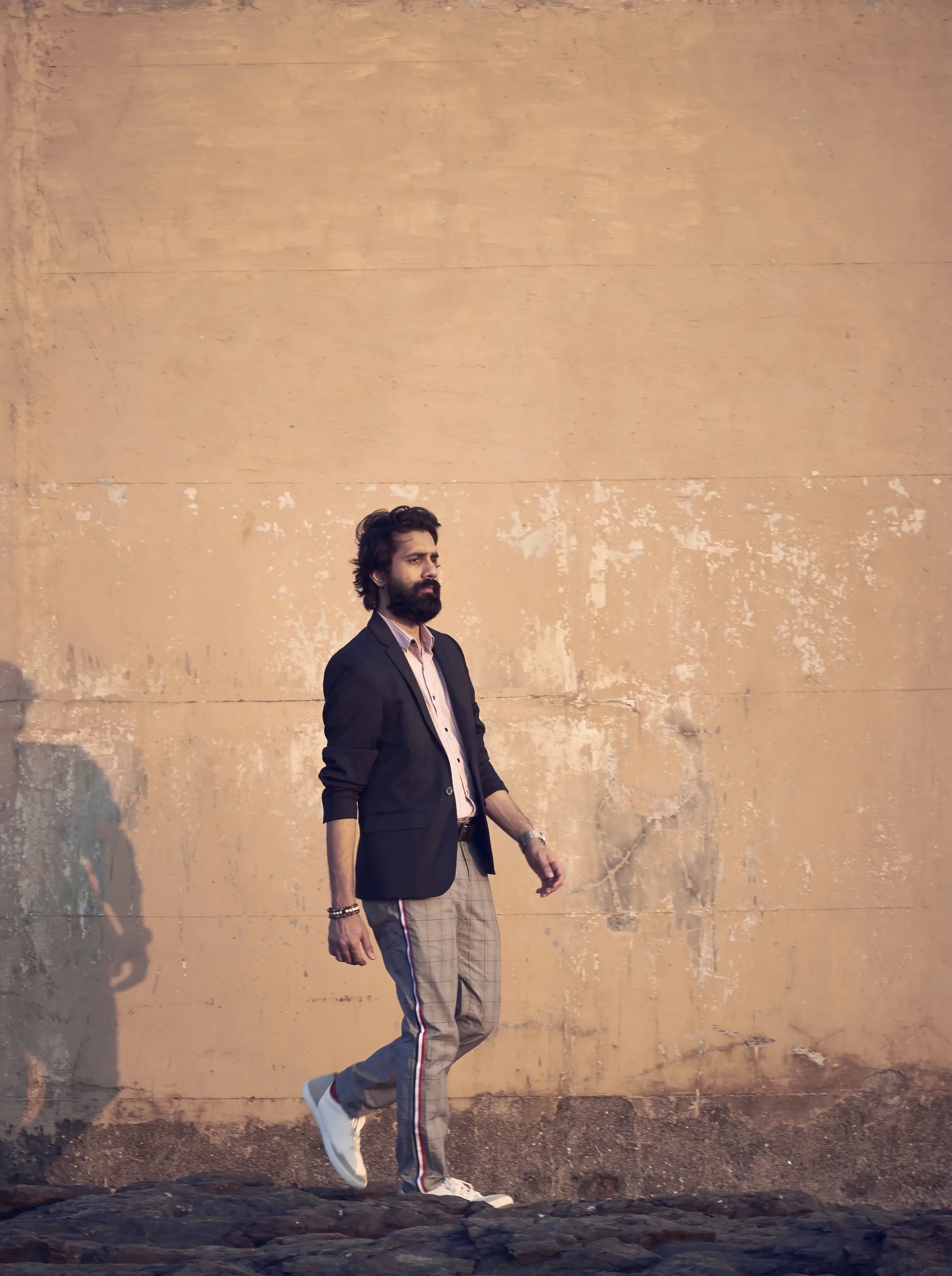 A man with dark hair and a beard, dressed in a black blazer, light pink shirt, and checked pants with red and white stripes, walking on a rocky surface in front of a large beige wall.